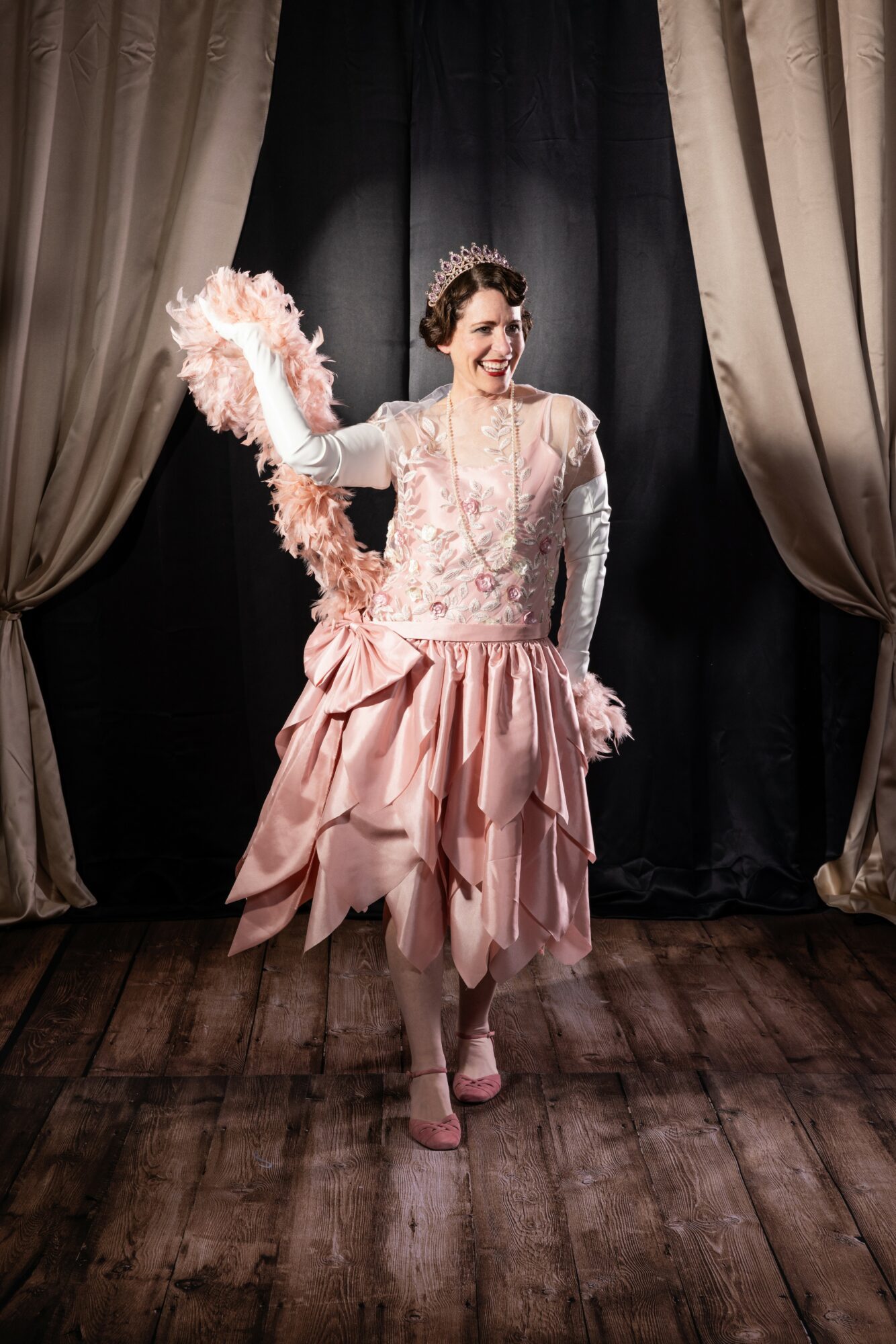 Woman in elegant pink and white dress with feathered accessory, smiling on stage with curtains behind her.