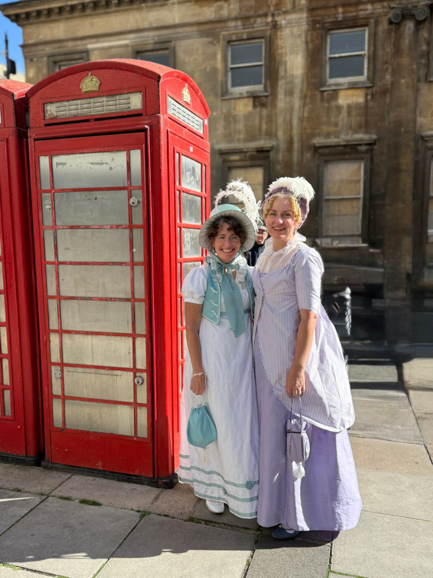 Two women in historical costumes stand next to a red British telephone booth on a city street, smiling at the camera.