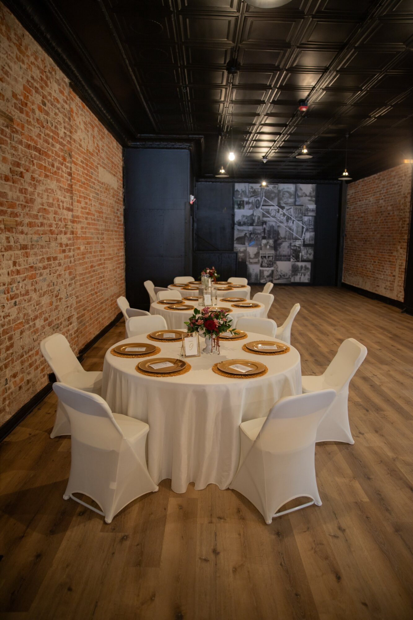 Round table with white chairs, set with plates, glasses, and a floral centerpiece, in a room with brick and dark walls.