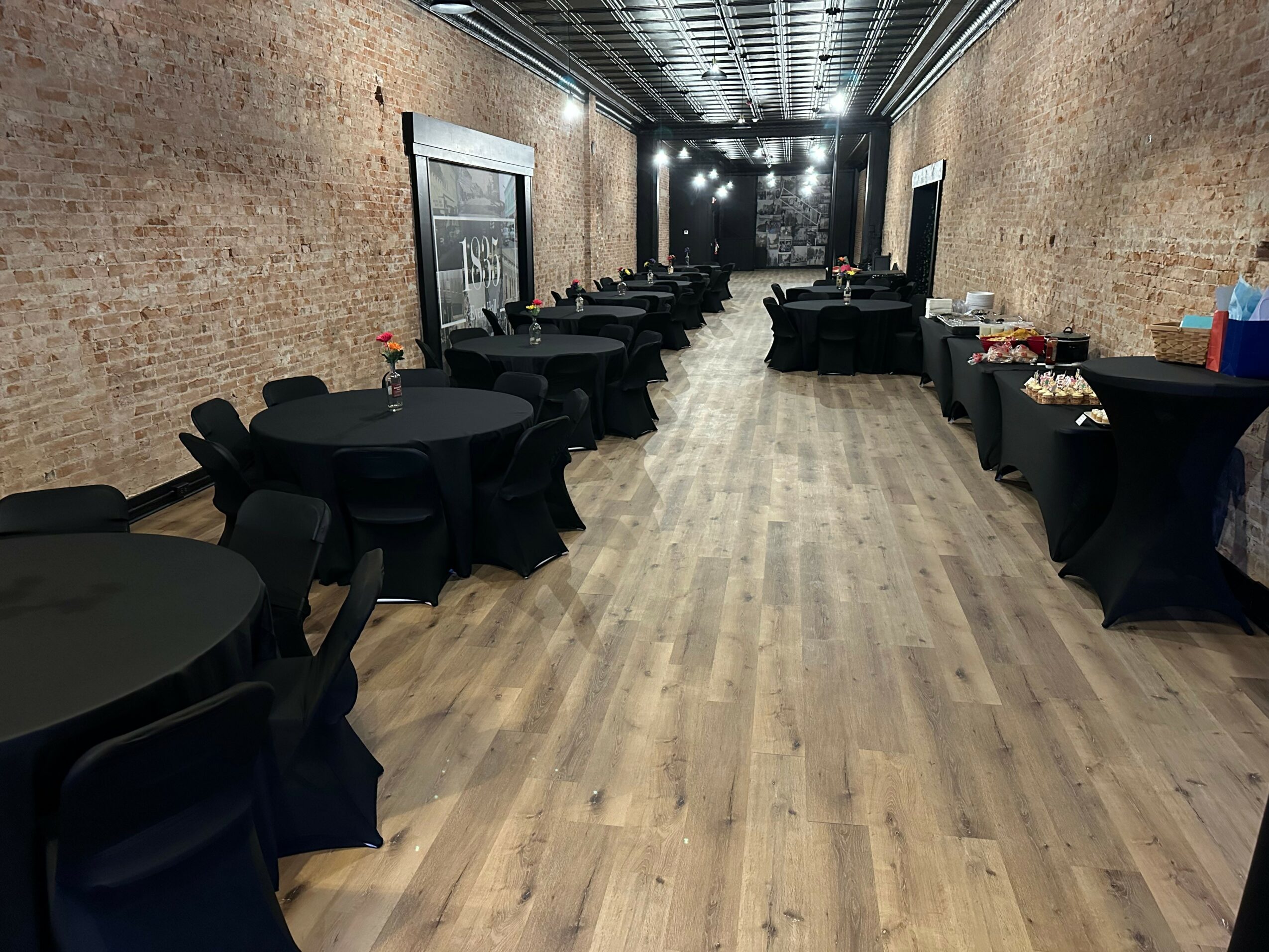 Long room with brick walls, wooden floor, black round tables, and chairs, set for an event with food and drinks.