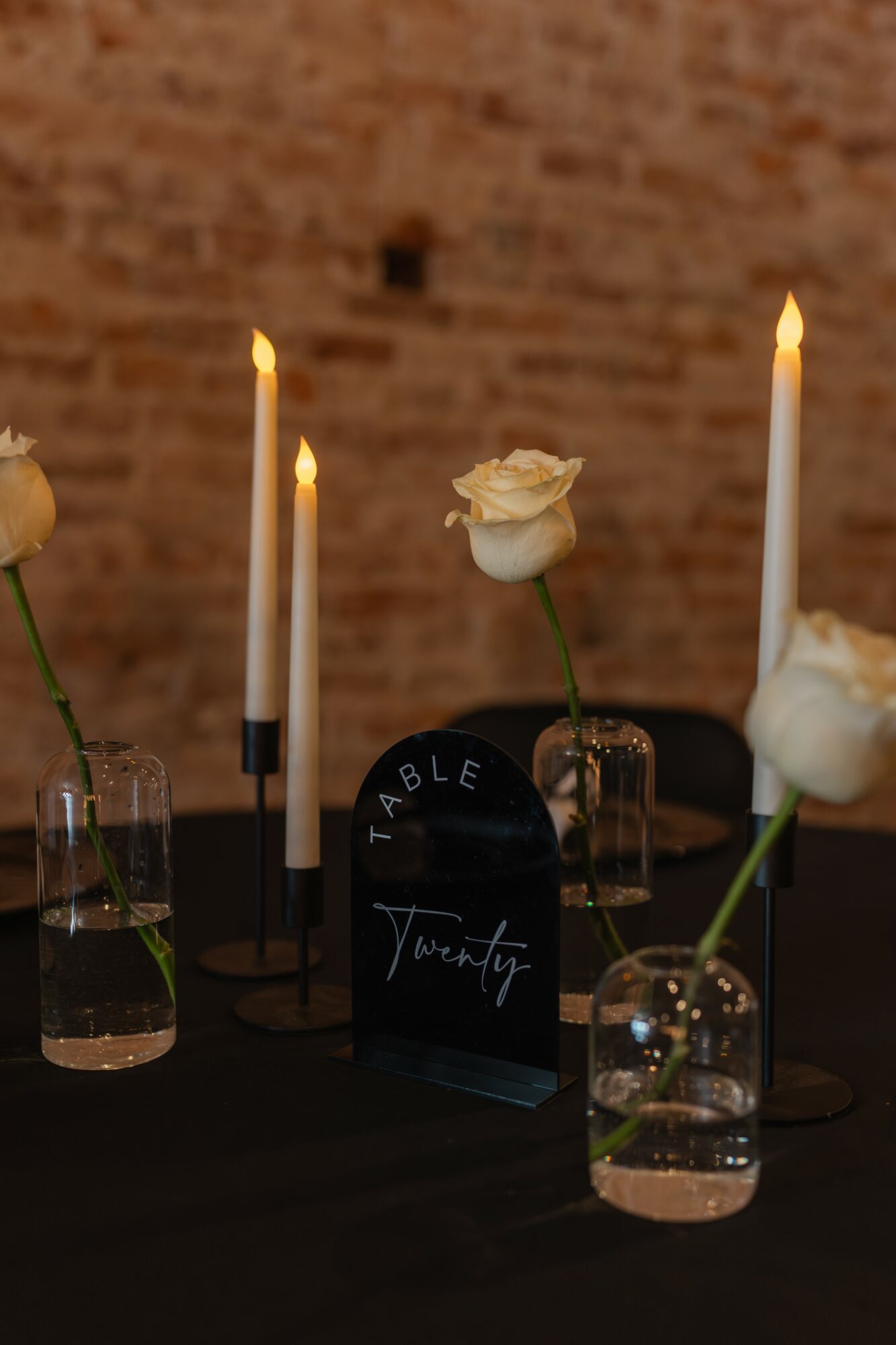Table setting with white roses in vases, tall candles, and a black table number sign, against a brick wall background.