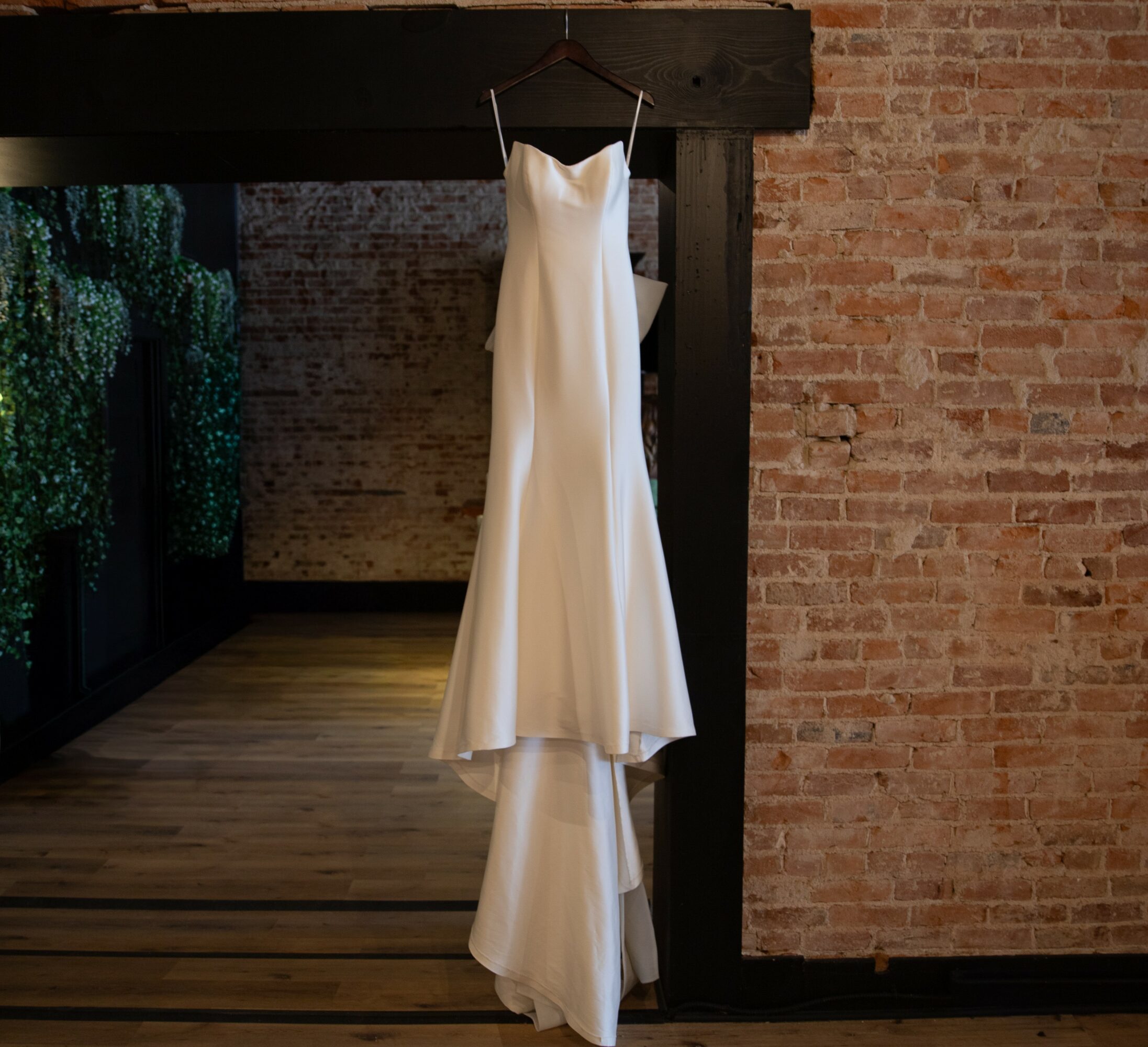 White wedding dress hanging on a hook against a brick wall, with wooden floor and dark wooden beam above.