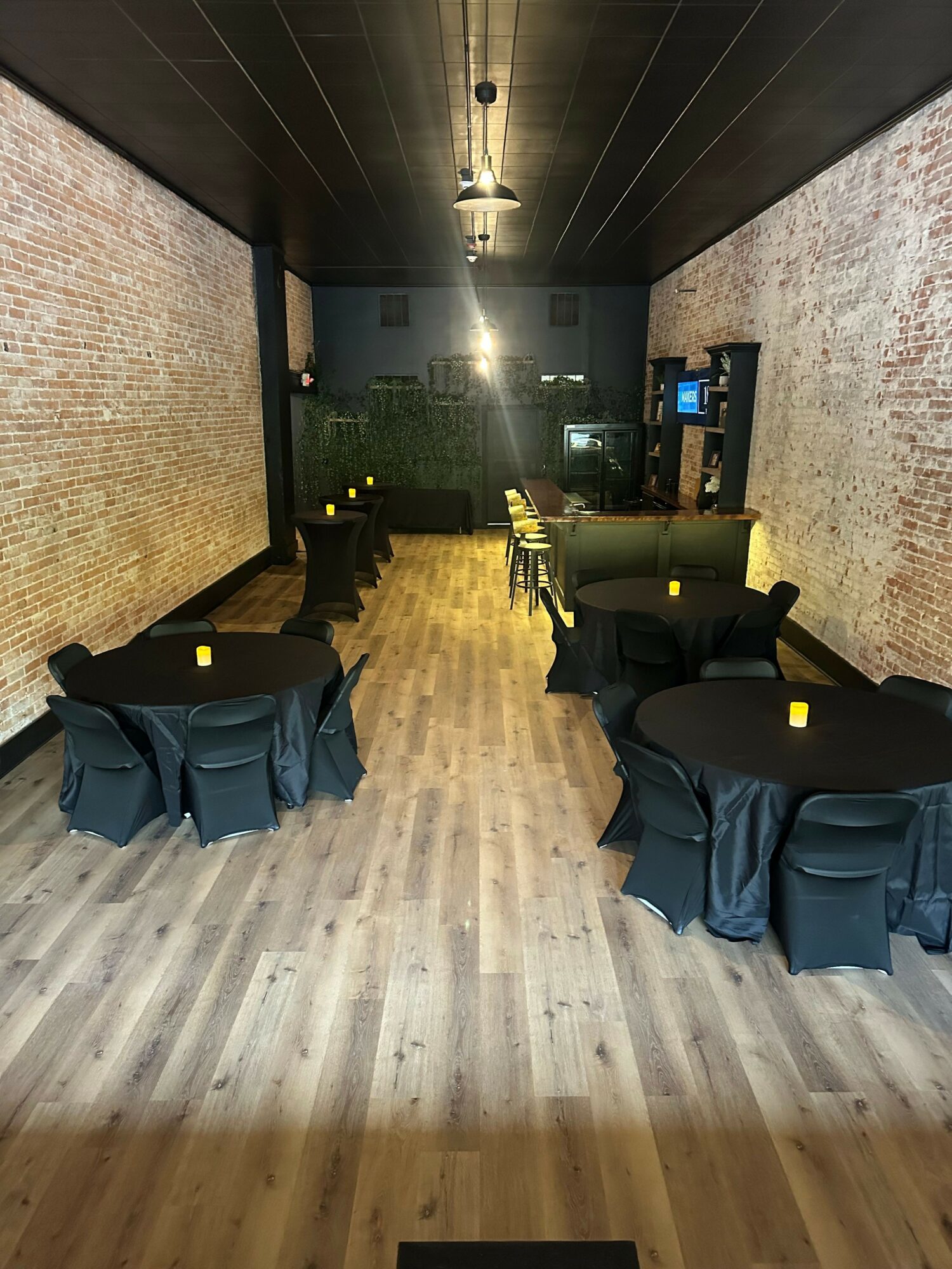 Indoor lounge with chairs, tables, and a brick wall, illuminated by ceiling lights, with wooden flooring.
