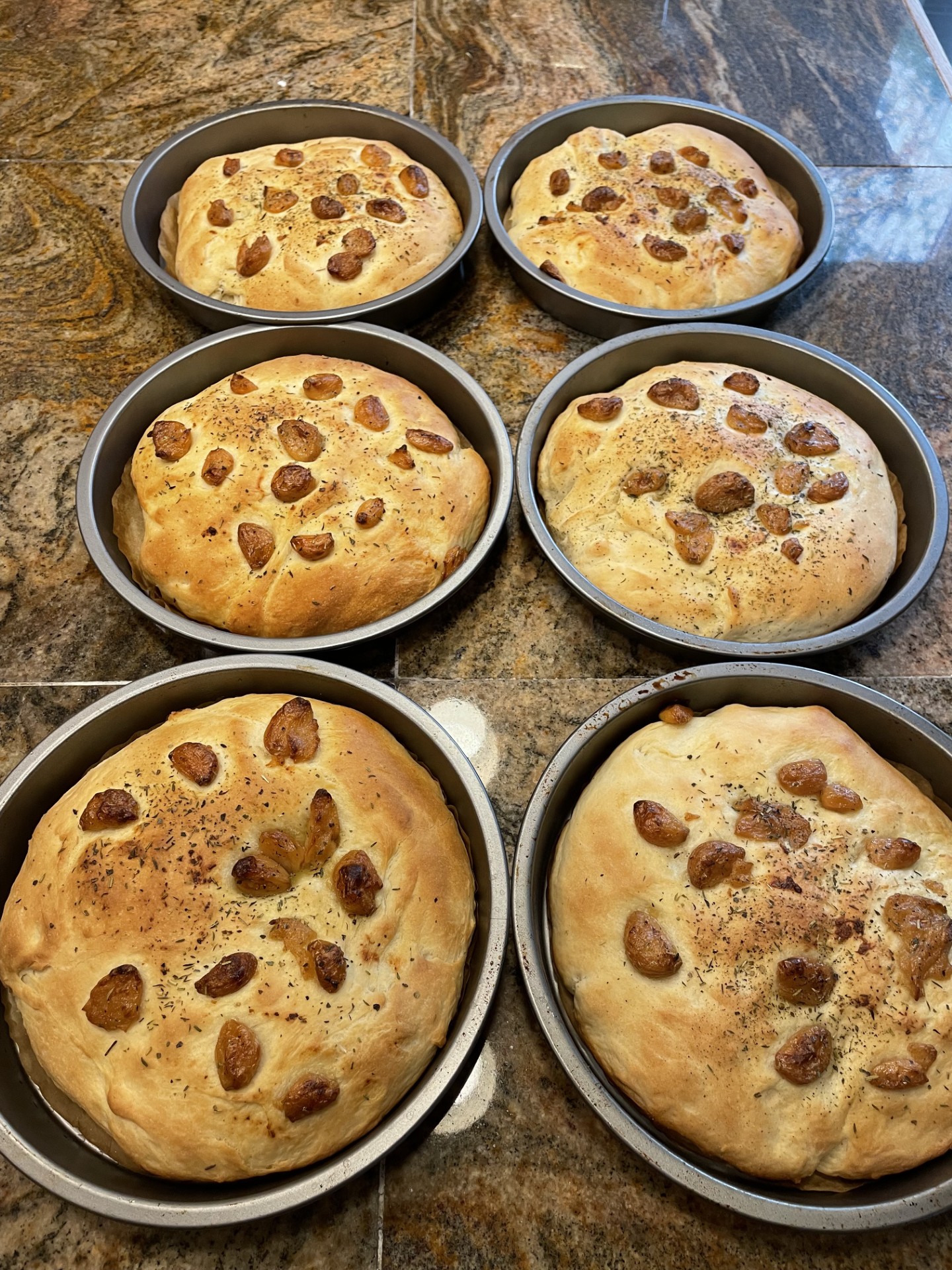 Six round baking pans with baked chocolate chip cookies on a dark marble countertop.