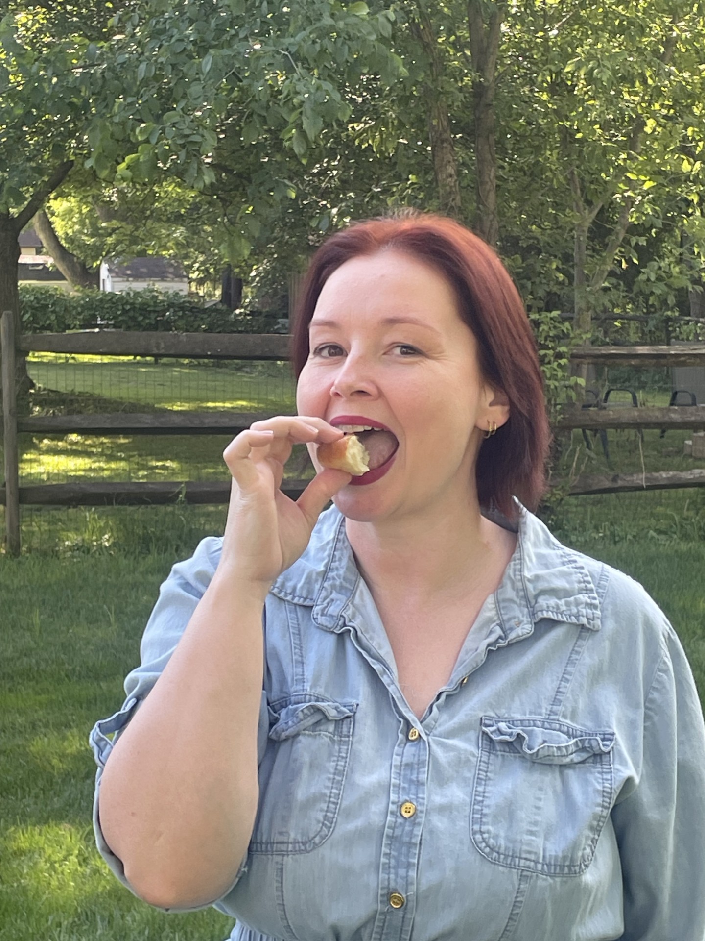 Woman with shoulder-length hair eating a snack outdoors, trees and a fence in the background.