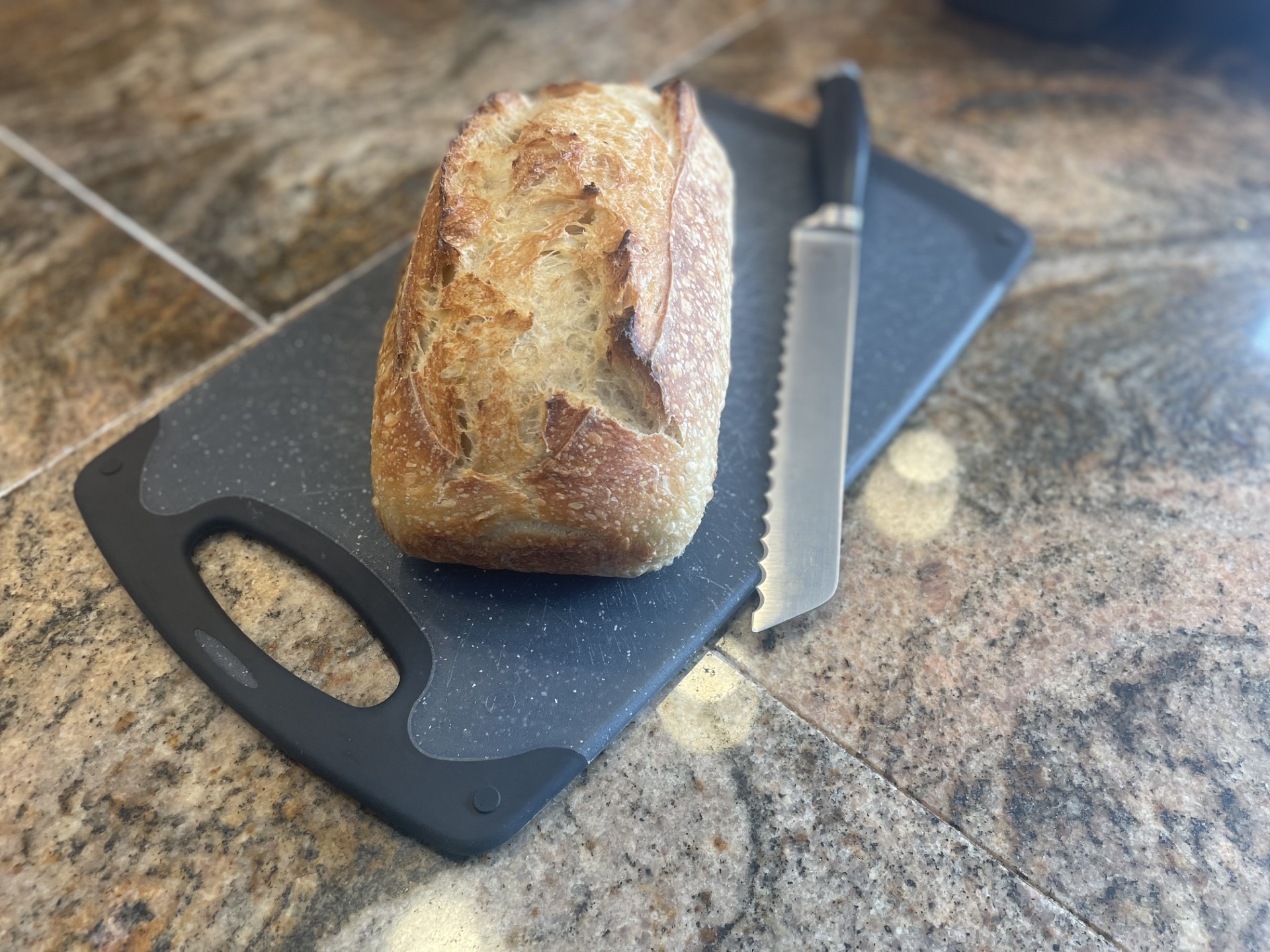 A loaf of bread on a black cutting board with a serrated knife, on a granite countertop.