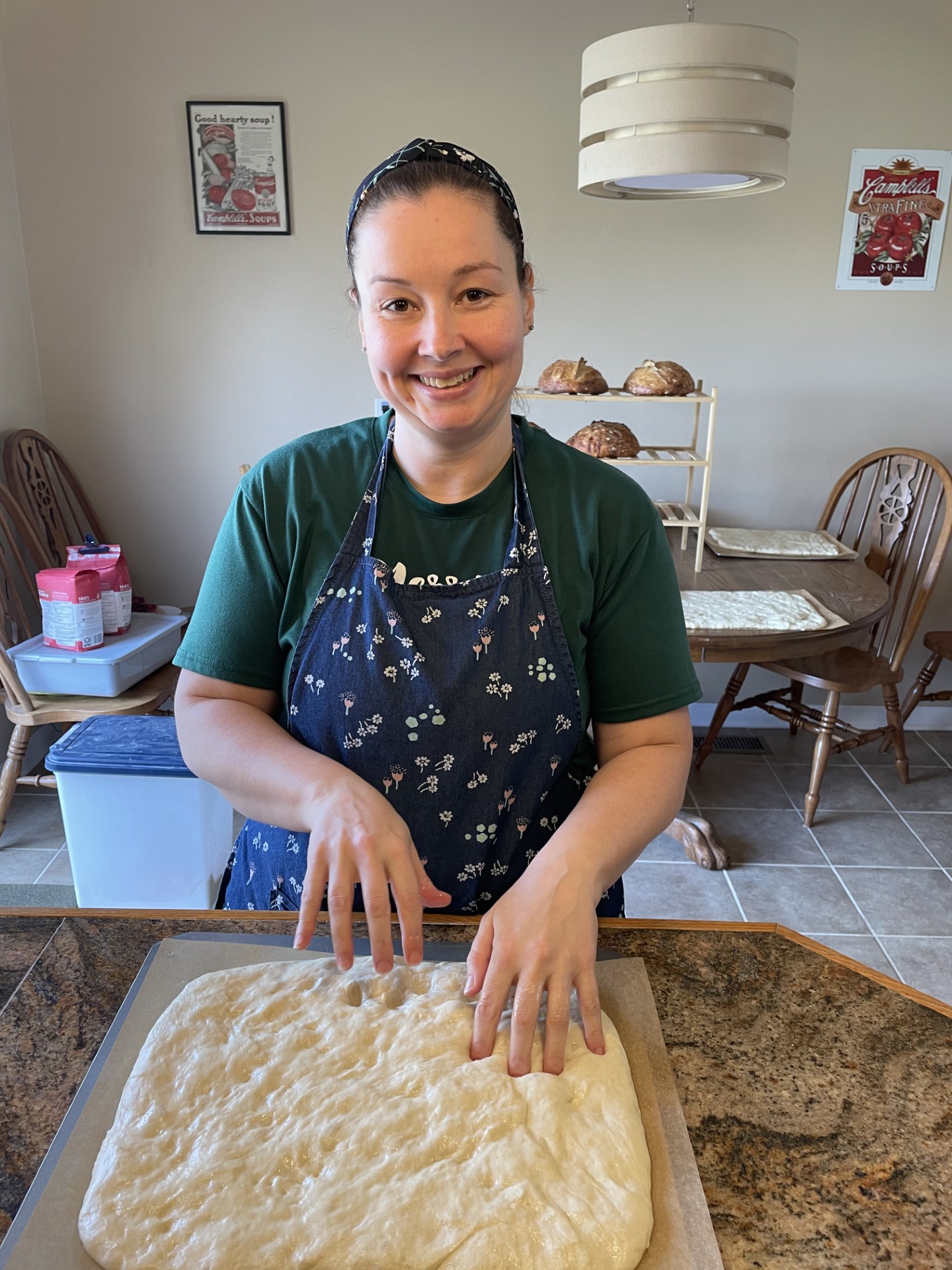 Smiling woman in a green shirt and apron preparing dough on a baking tray in a kitchen.