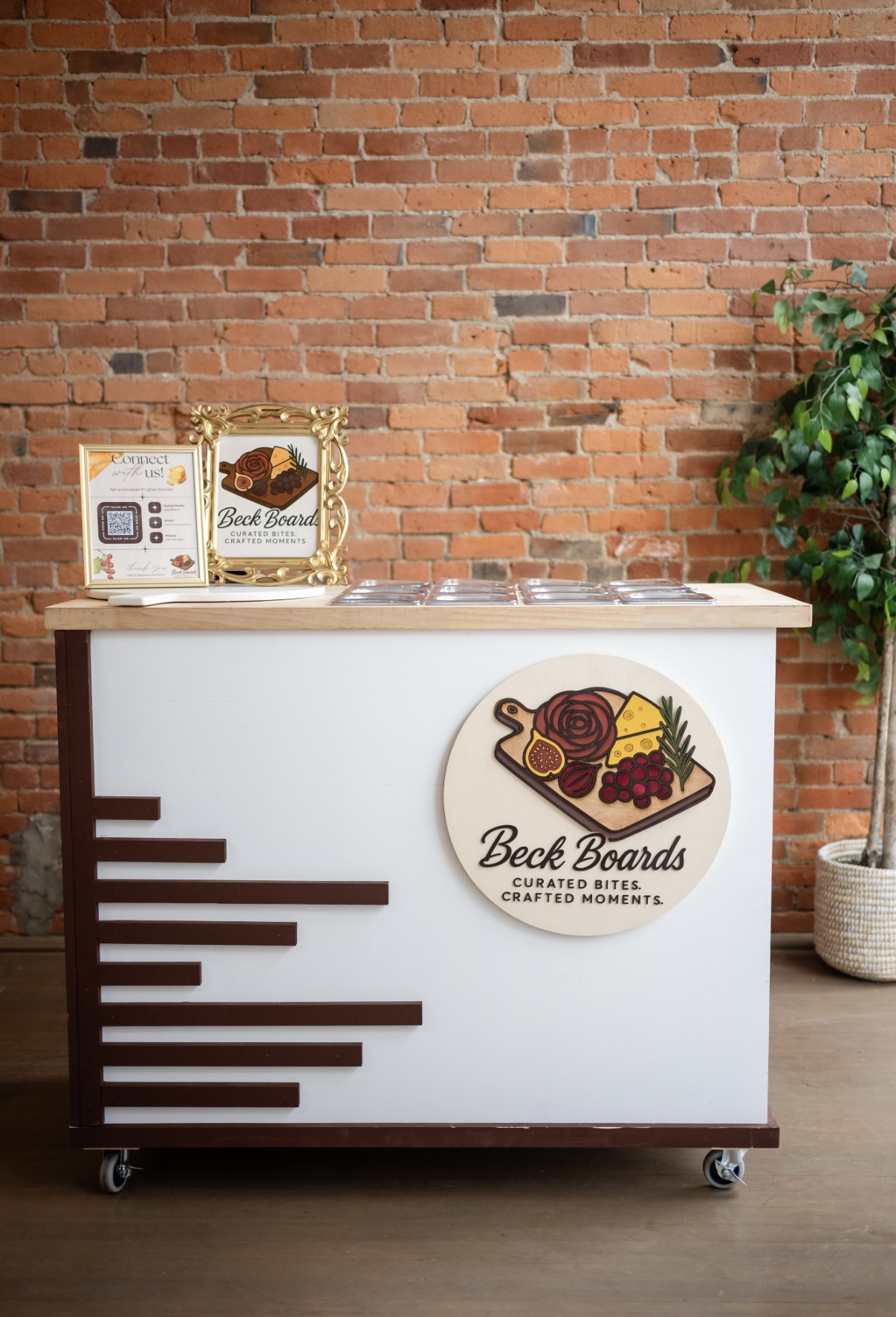 White reception desk with logo, brick wall background, potted plant, and awards on top, on a wooden floor.