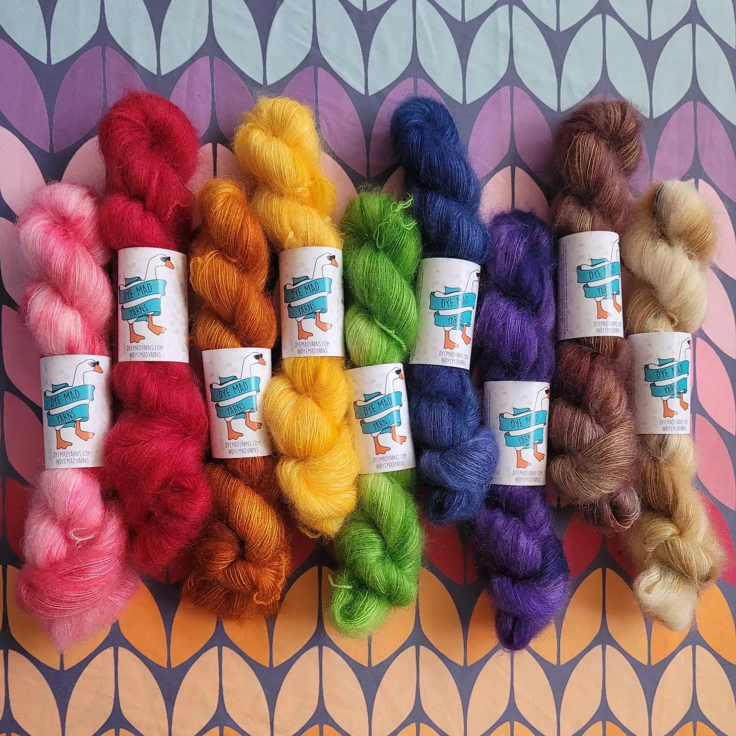 Six skeins of colorful yarn arranged in a row on a patterned background, from pink to beige, with labels attached.