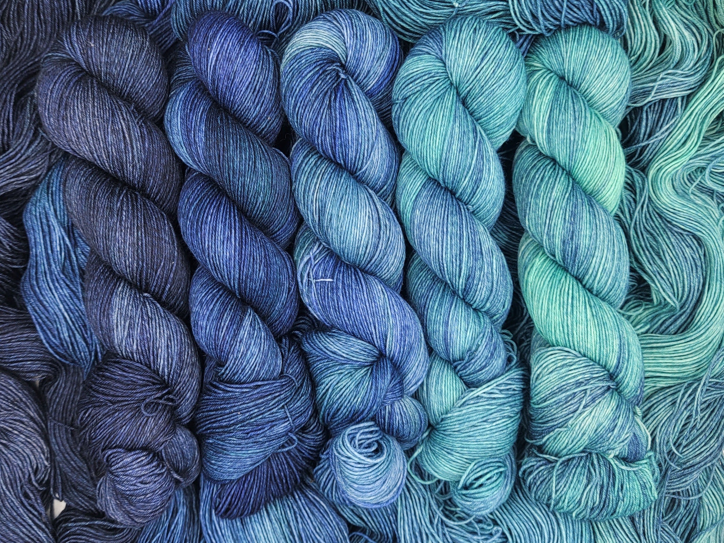Multiple skeins of yarn in shades of blue and green, arranged side by side, twisted into hanks.