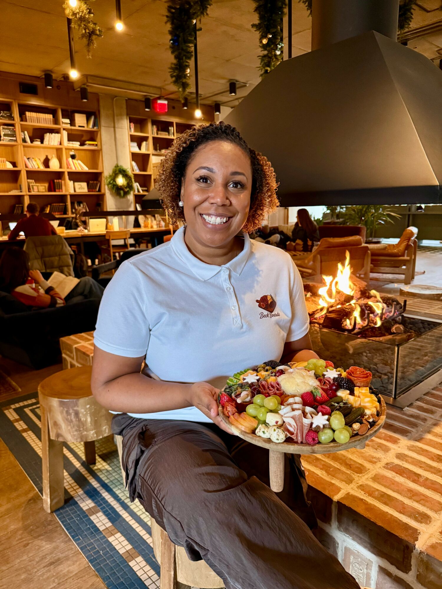 Smiling woman holding a large platter of colorful food in a cozy restaurant with a fireplace.