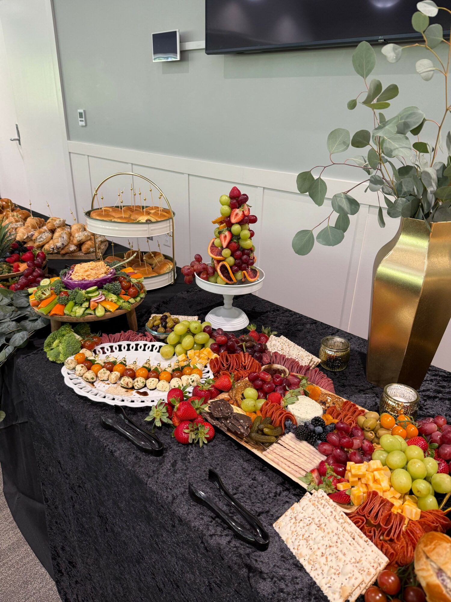 Table with fruit, cake, and snacks, decorated with flowers and a black tablecloth, in a modern room.