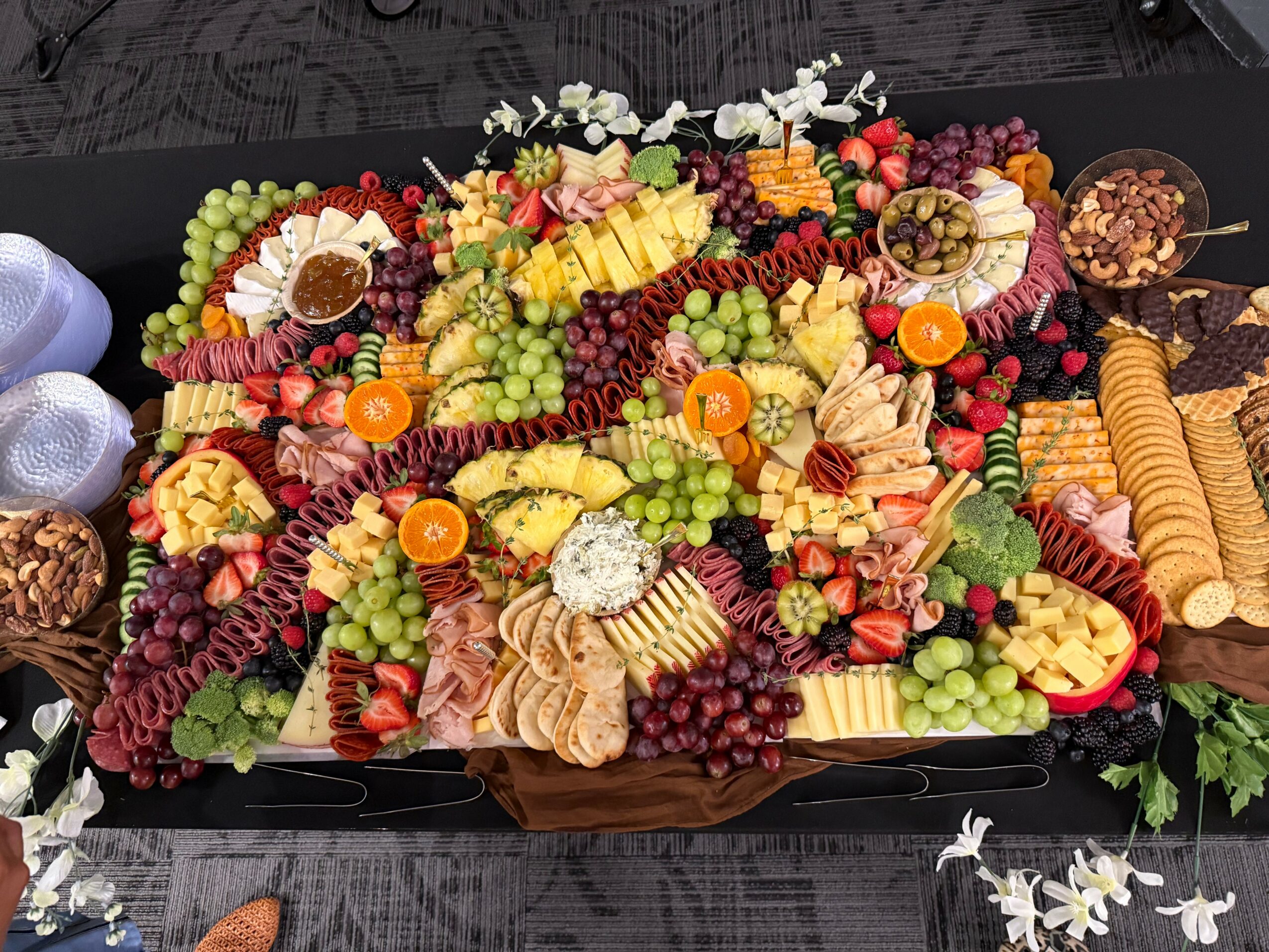Assorted fruit platter with grapes, pineapple, and other fruits, arranged on a black tray with crackers and nuts, viewed from above.