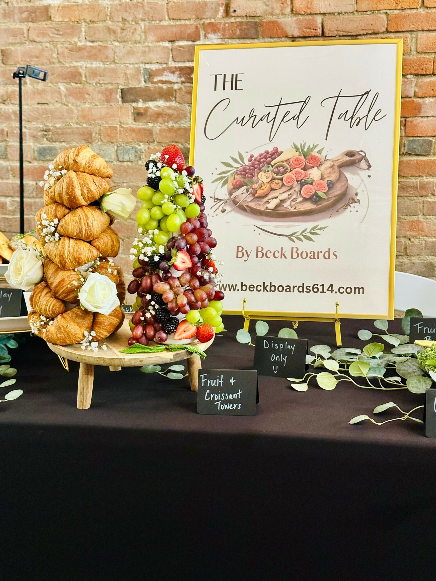 Display of croissants, grapes, and berries on a table with a framed sign reading 'The Crafted Table' and a brick wall background.