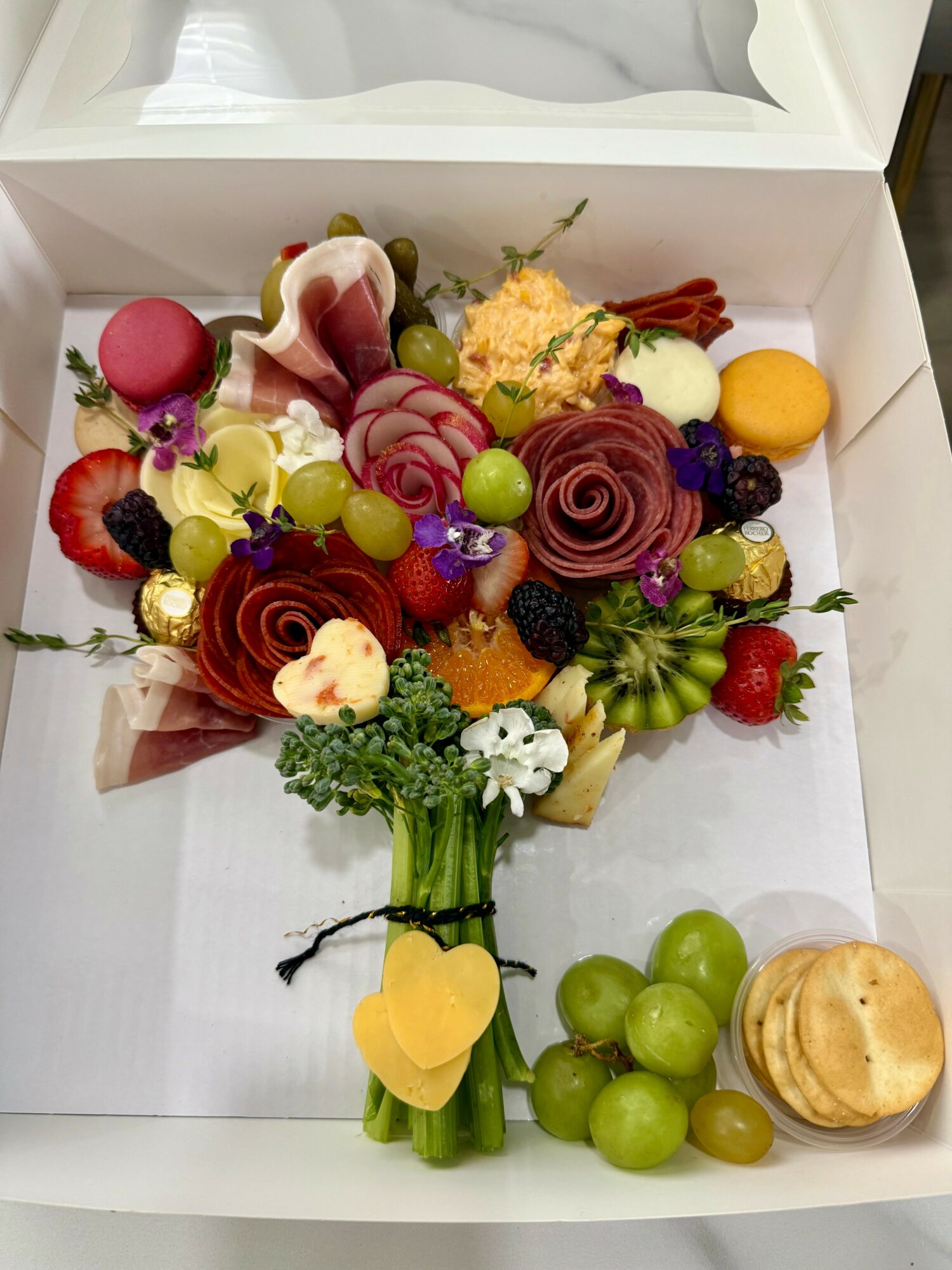 Colorful fruit and flower arrangement with grapes, strawberries, and decorative flowers in a white box.