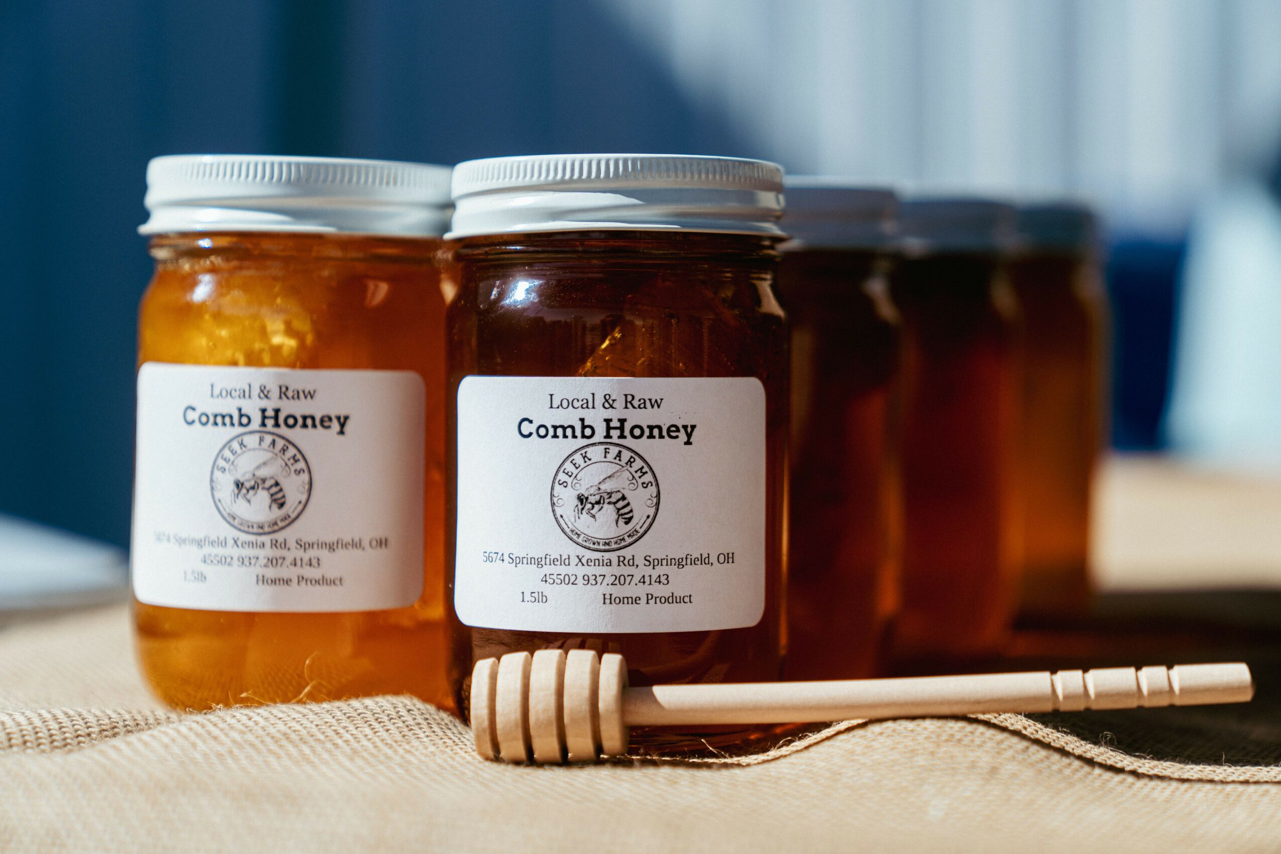 Three jars of comb honey with a honey dipper on a cloth surface, background of blue curtains, natural lighting.