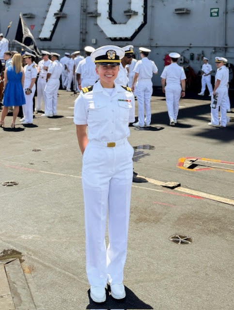 Woman in white naval uniform smiling, standing outdoors with a group of people in similar uniforms and a ship in background.
