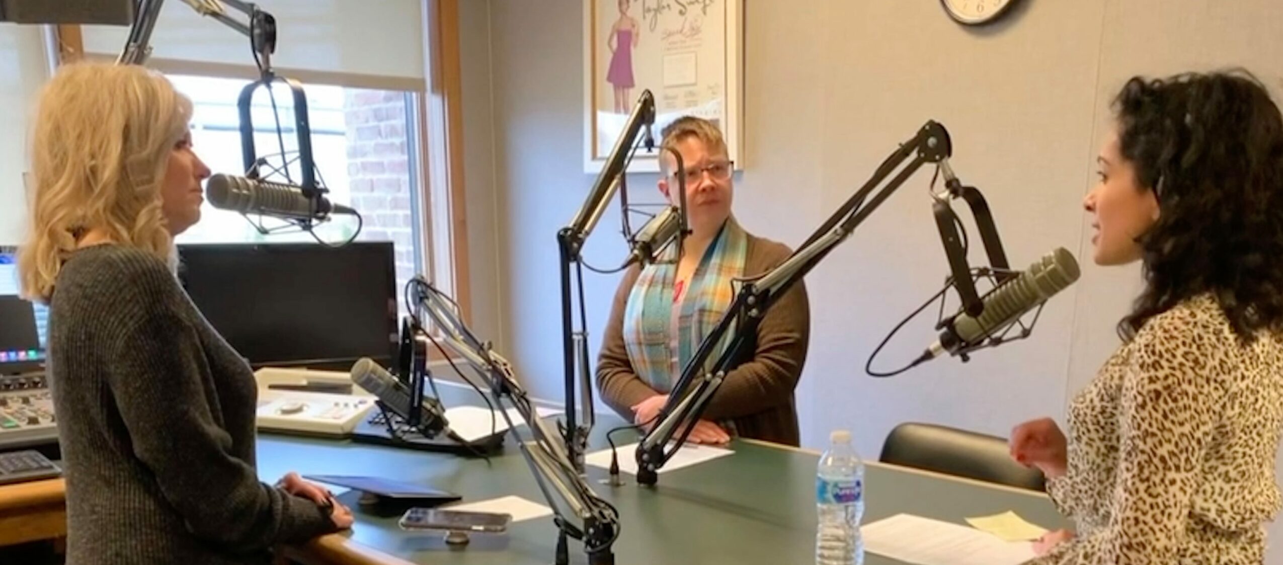 Three women sit at a table in a radio studio with microphones, discussing. One woman has a water bottle in front of her.