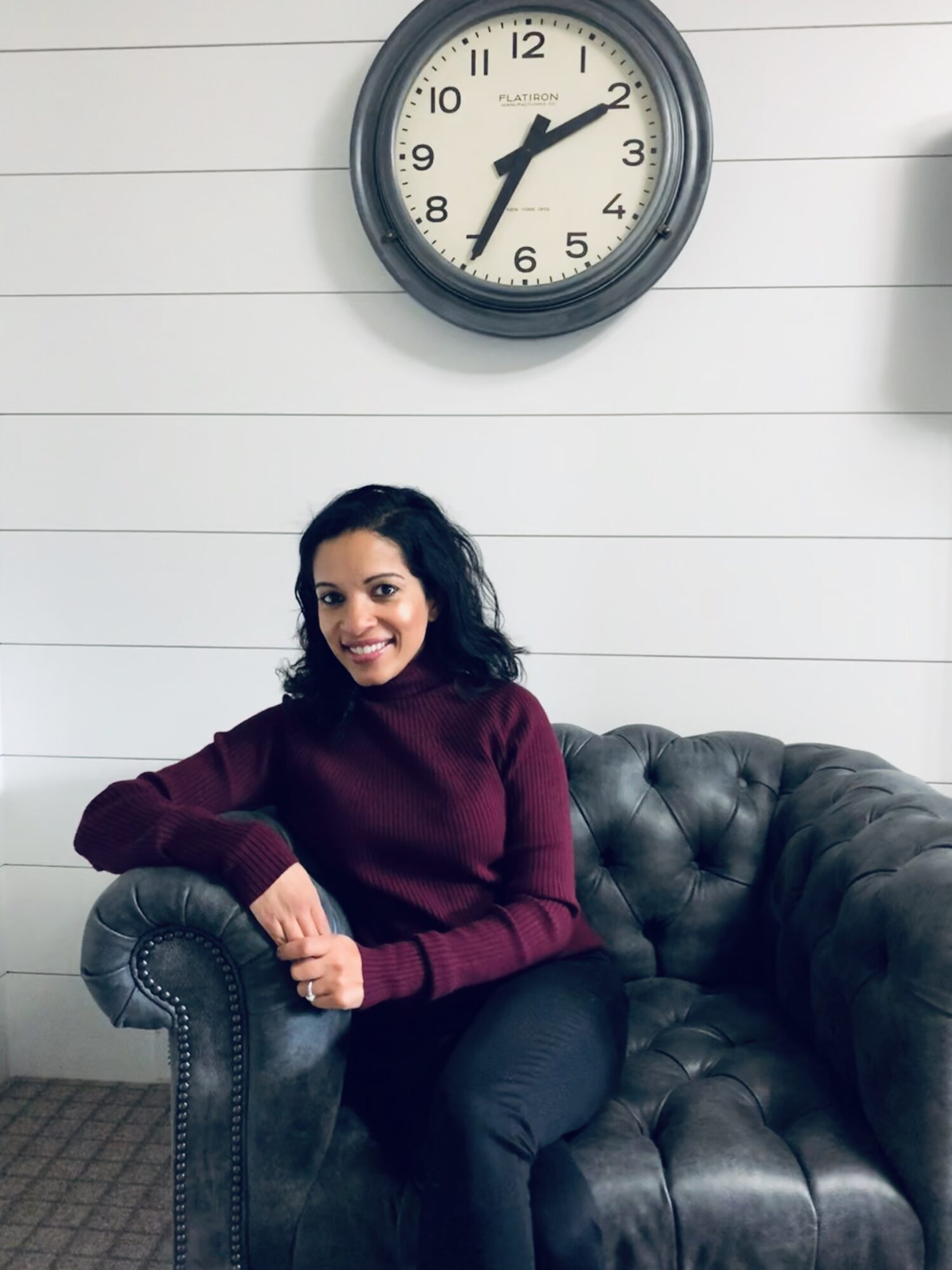 Woman sitting on a black leather couch, smiling, with a clock showing 2:10 on the wall behind her.