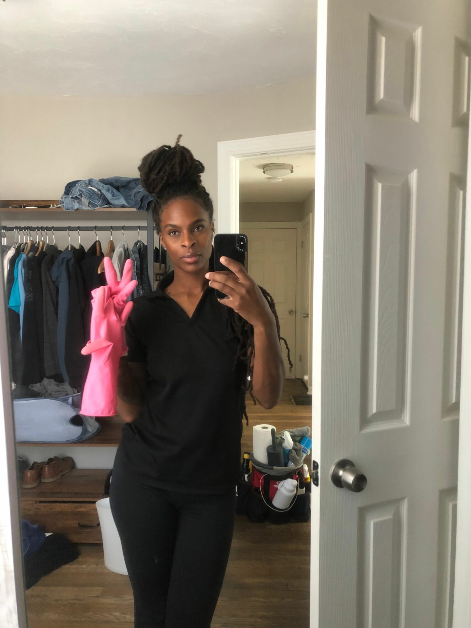 Person taking a mirror selfie in a room with clothes hanging and a door in the background.