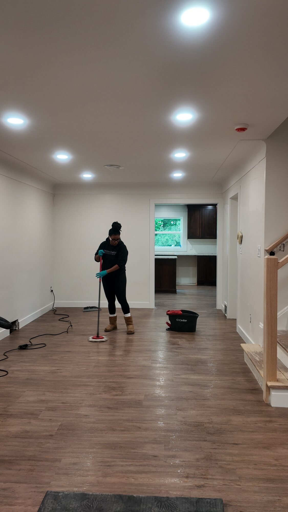 Person mopping wooden floor in a spacious, well-lit room with a kitchen area in the background.