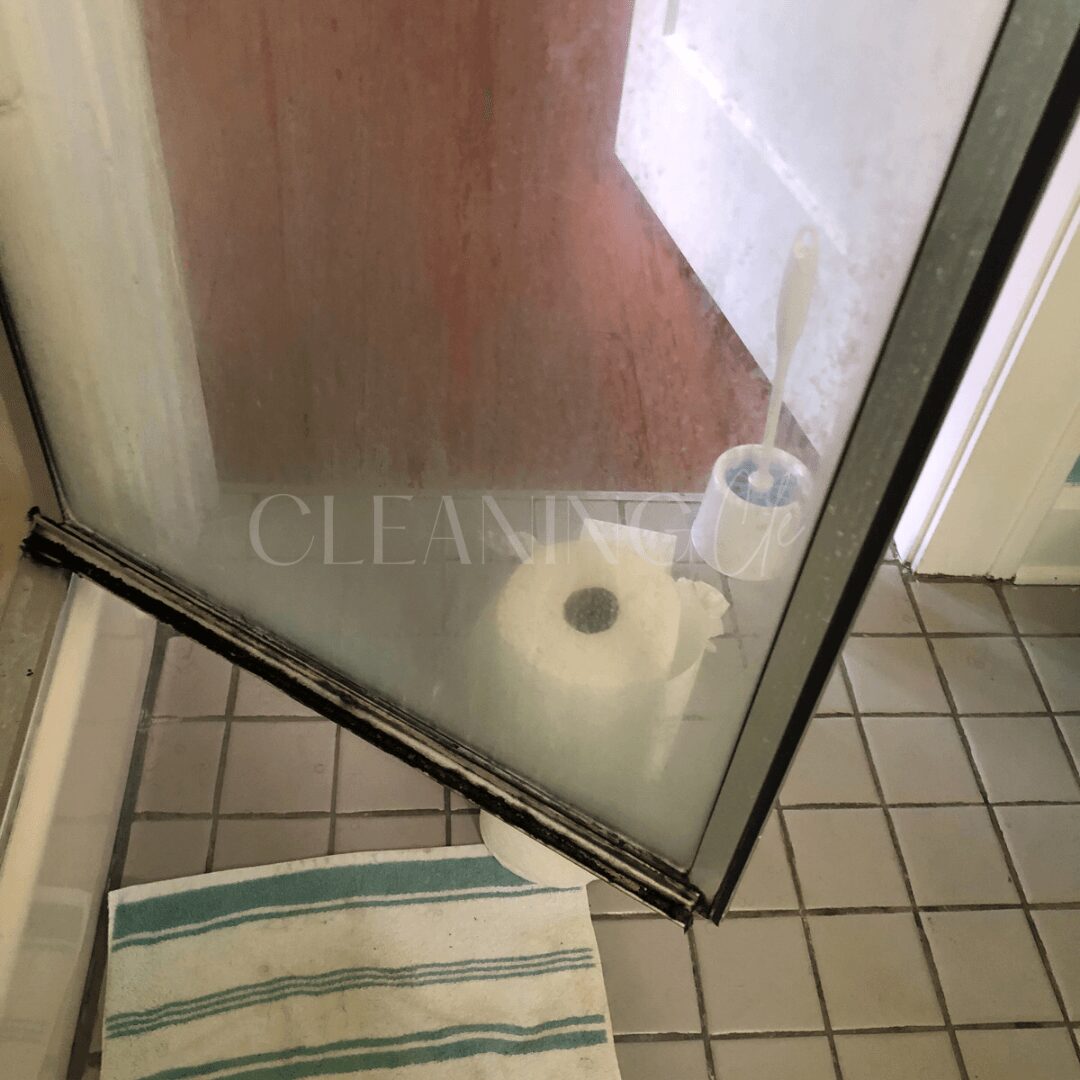 View through a glass shower door showing a roll of toilet paper and a tiled floor.