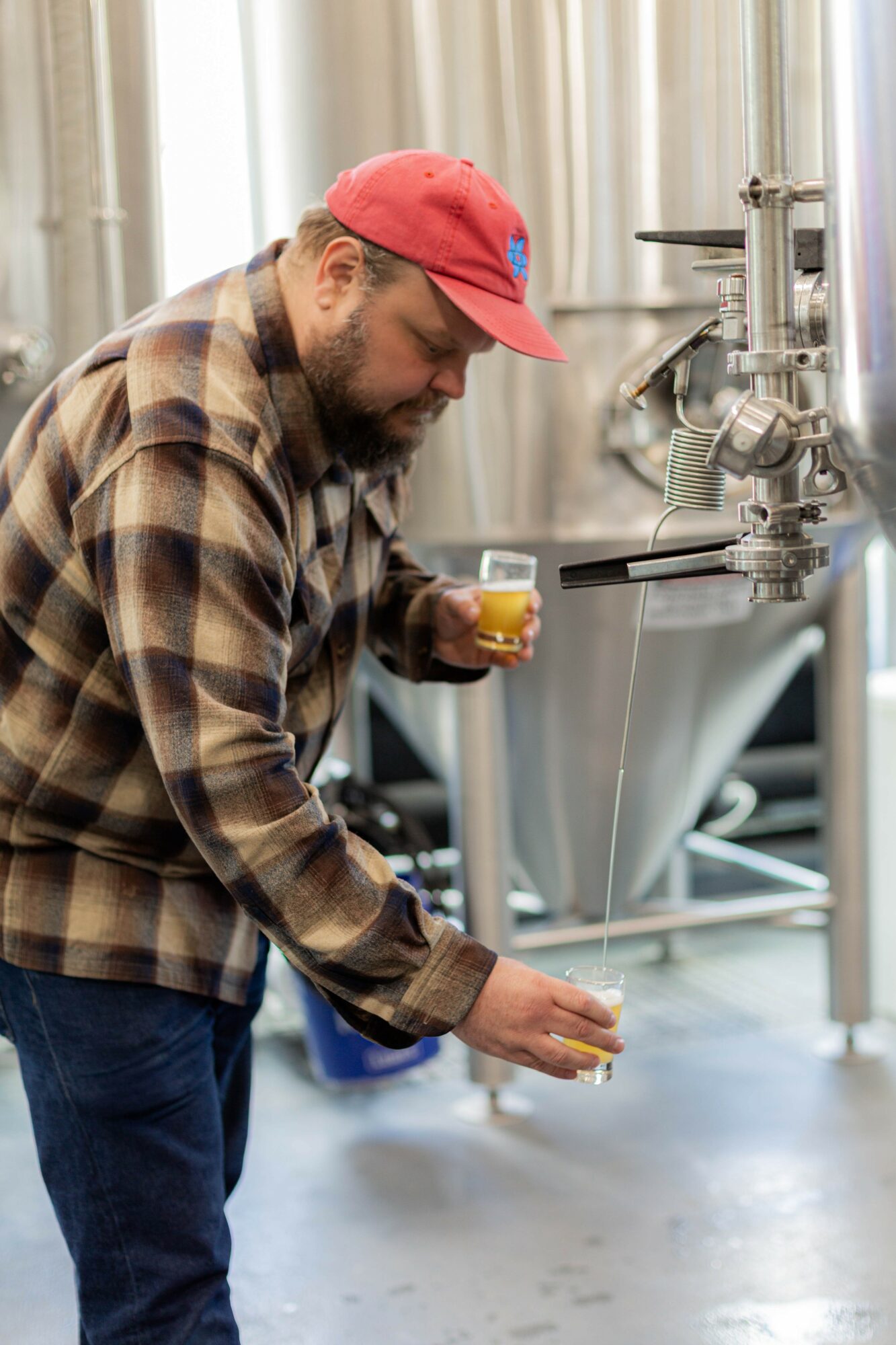Man in plaid shirt and red cap pours beer from tap into glass in brewery setting.