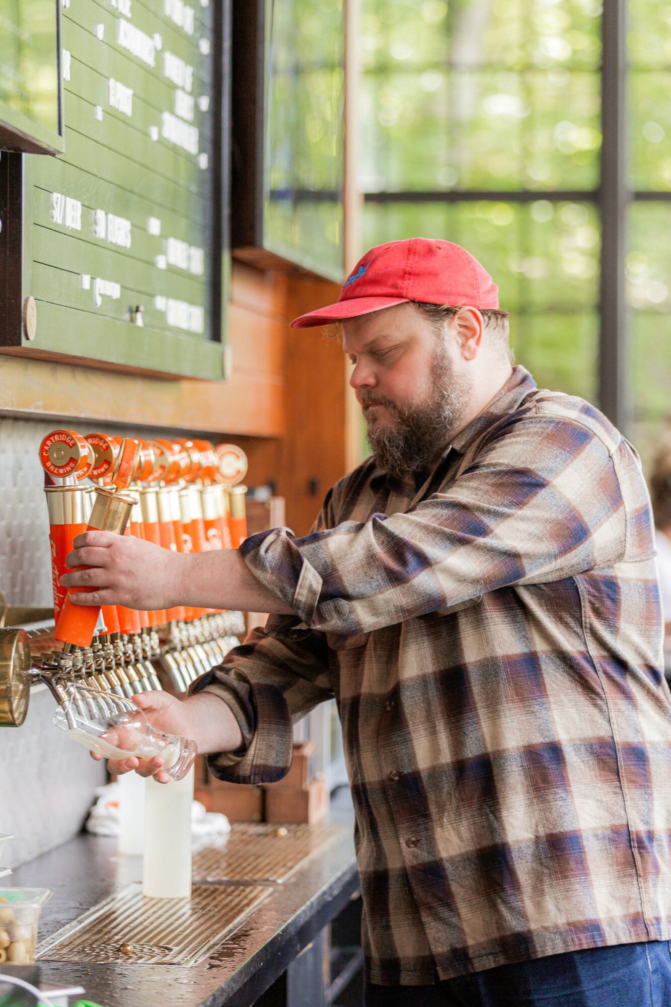 Man with a beard in a plaid shirt and red cap fills a glass at a bar with beer taps, in a bright setting.