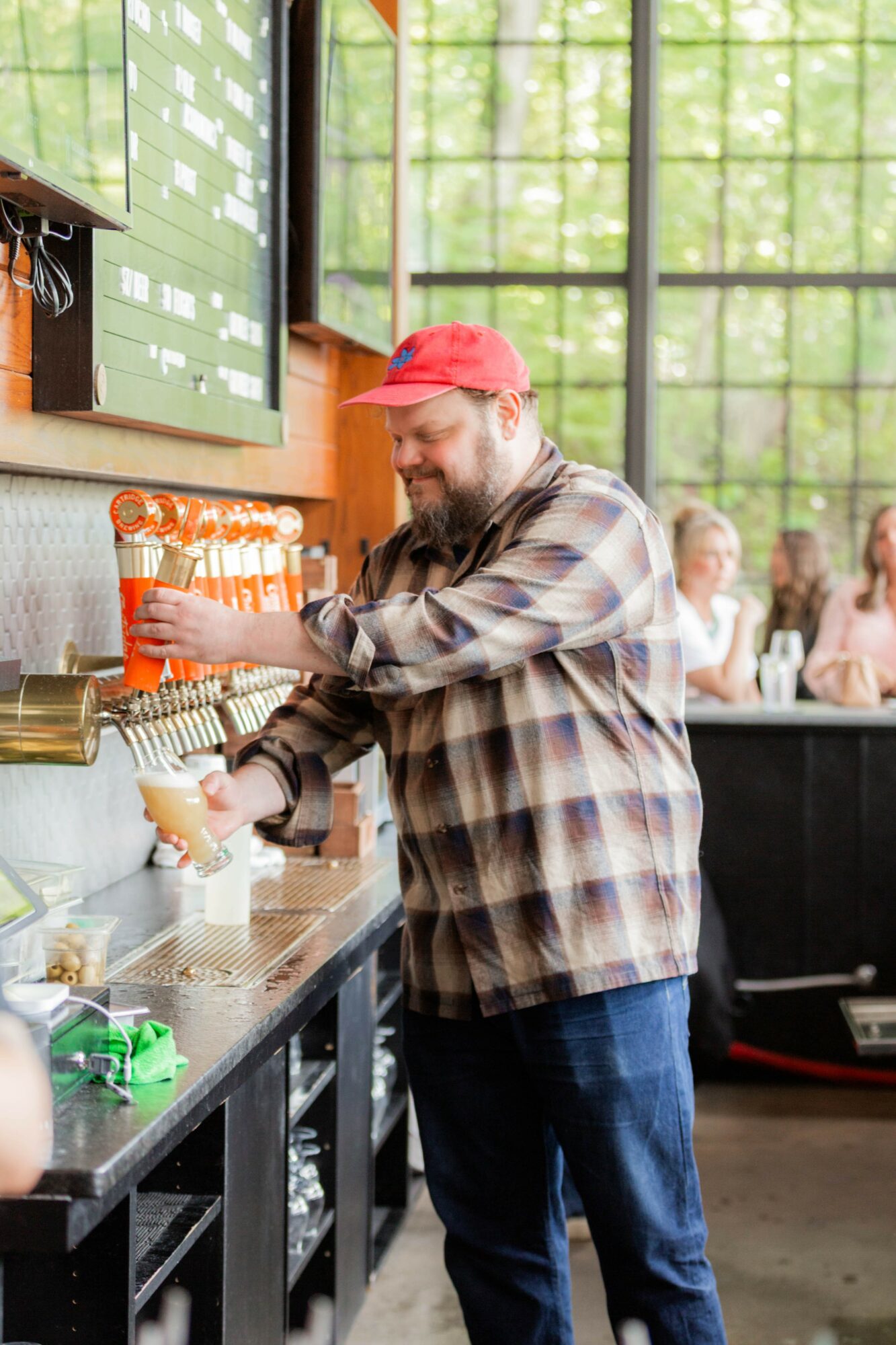 Man with a beard in a plaid shirt and red cap pours a drink at a bar counter inside a cafe with large windows.