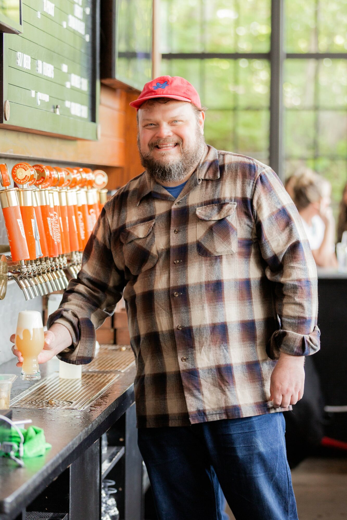 Man with a beard and red cap pouring beer at a bar, smiling, in a bright indoor setting.