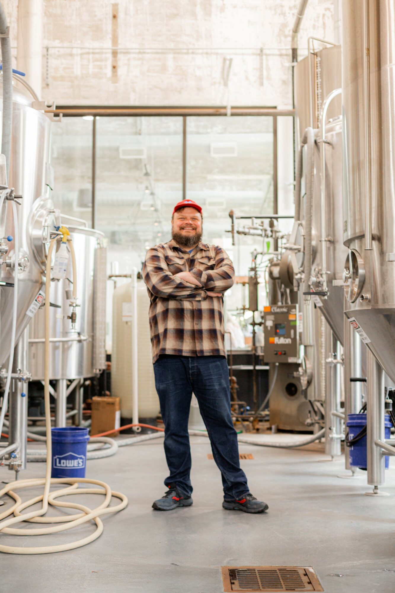 Man with red hair and beard wearing plaid shirt and jeans standing with arms crossed in industrial setting with tanks and pipes