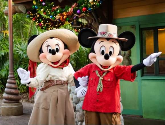 Mickey Mouse and Minnie Mouse dressed in festive costumes, standing outdoors with a decorated Christmas tree in the background.