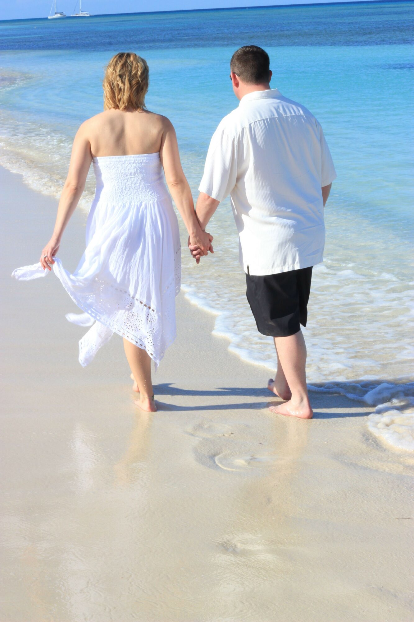 A woman in a white dress and a man in a white shirt and black shorts walk on the beach near the water's edge.