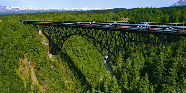 Train traveling on a bridge over a green forested valley with mountains in the background.