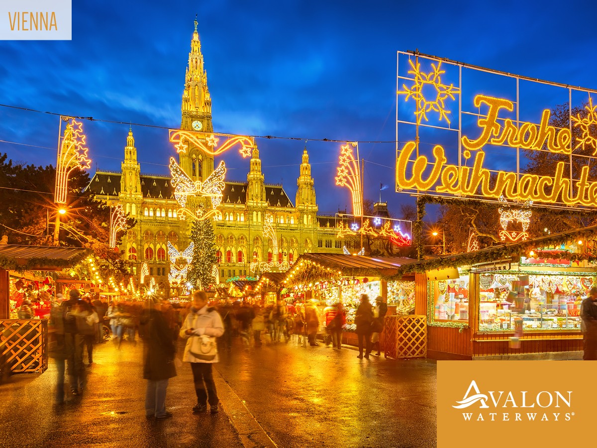 Night scene of a Christmas market in Vienna with illuminated historic buildings, festive lights, and market stalls, people walking around.