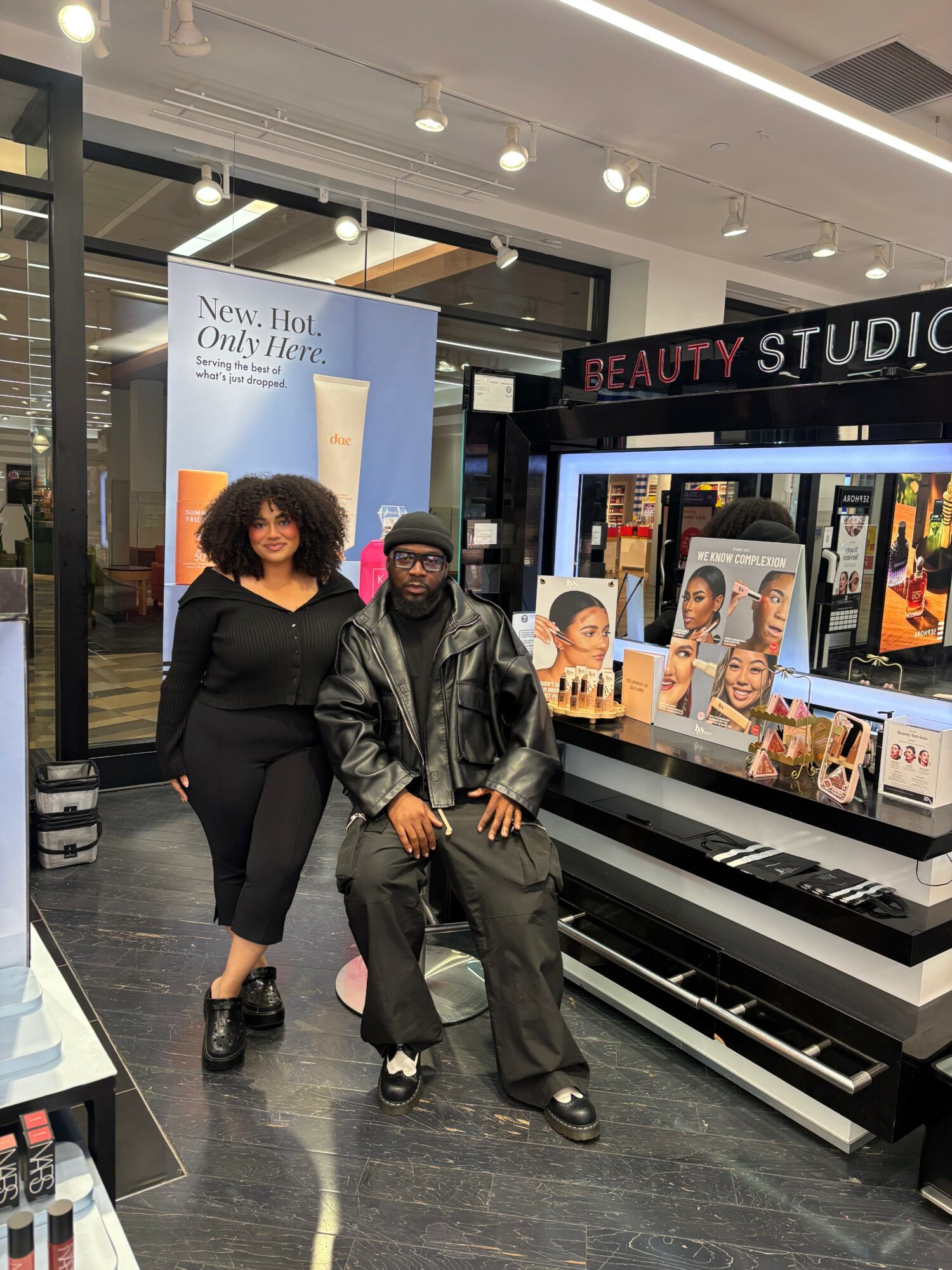 Two people sit inside a beauty store, one standing and one seated, with makeup displays and a sign in the background.