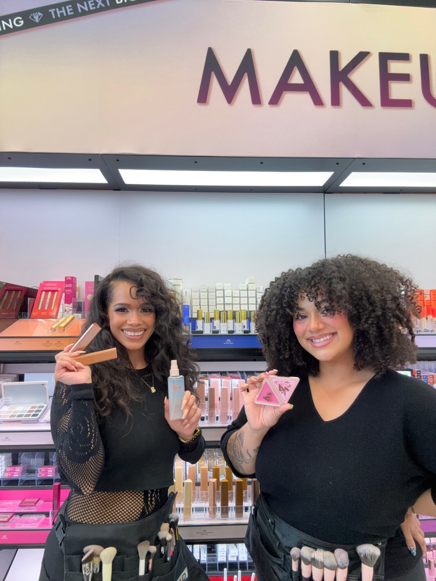 Two women with curly hair smiling in a makeup store, holding makeup products, shelves with beauty items behind them.