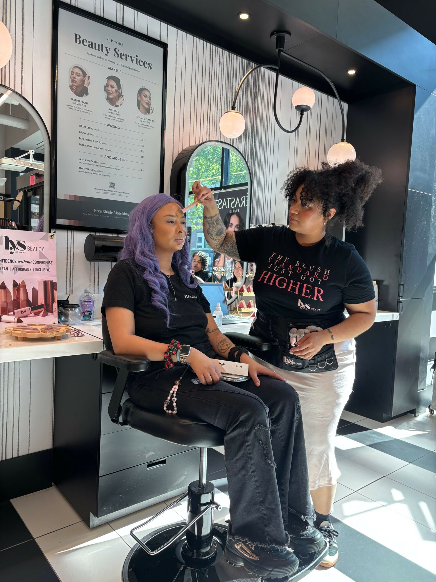 Two women in a salon, one sitting and the other standing, with a mirror and informational poster behind them.