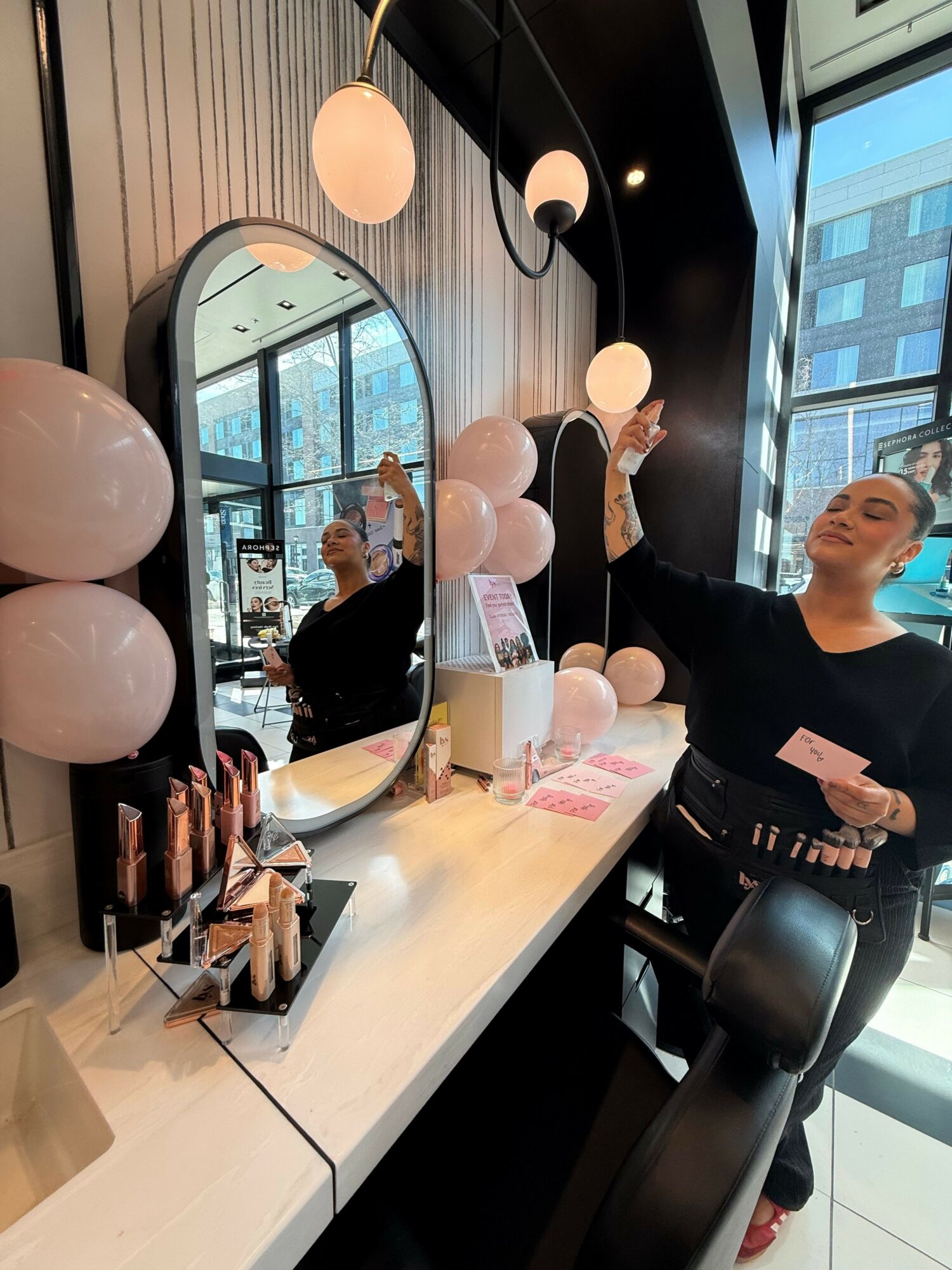 Woman standing at a makeup station with brushes, mirror, and balloons, in a modern room with large windows.