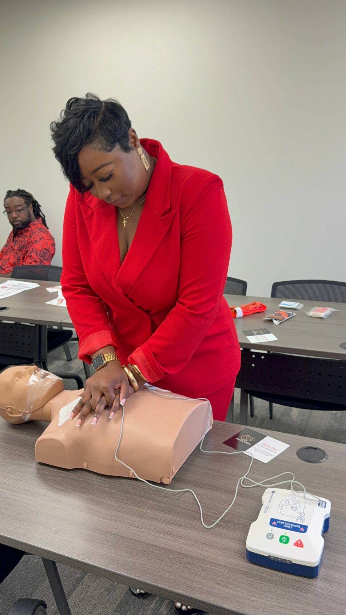 Woman in red suit practicing CPR on a mannequin in a classroom setting.