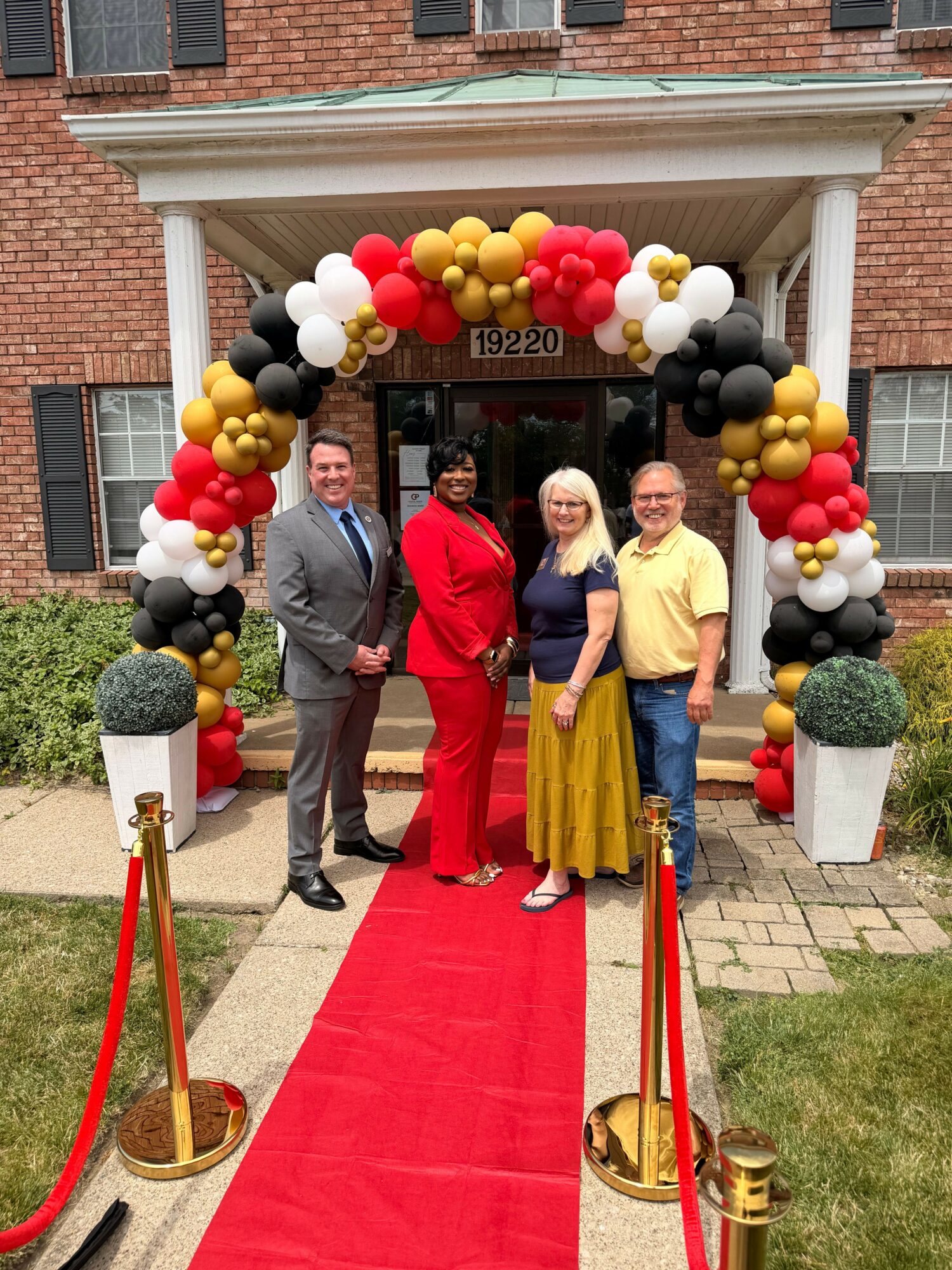 Three people standing on a red carpet outside a building, with a balloon arch in red, black, white, and gold colors, and a grassy area nearby.