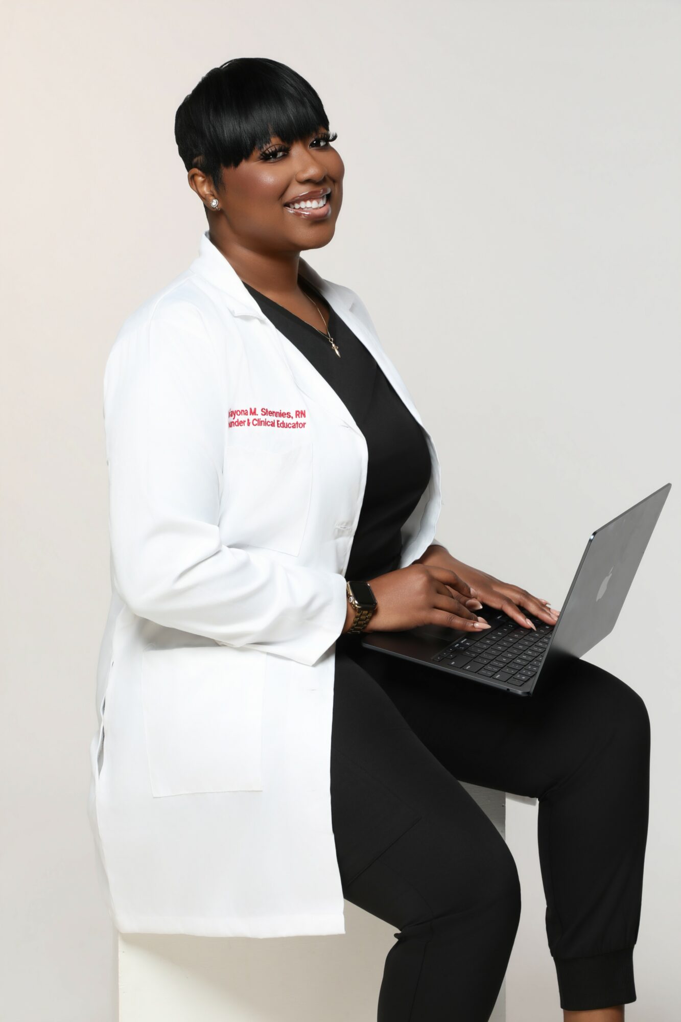 Woman in white lab coat sitting with a laptop, smiling, against a plain background.