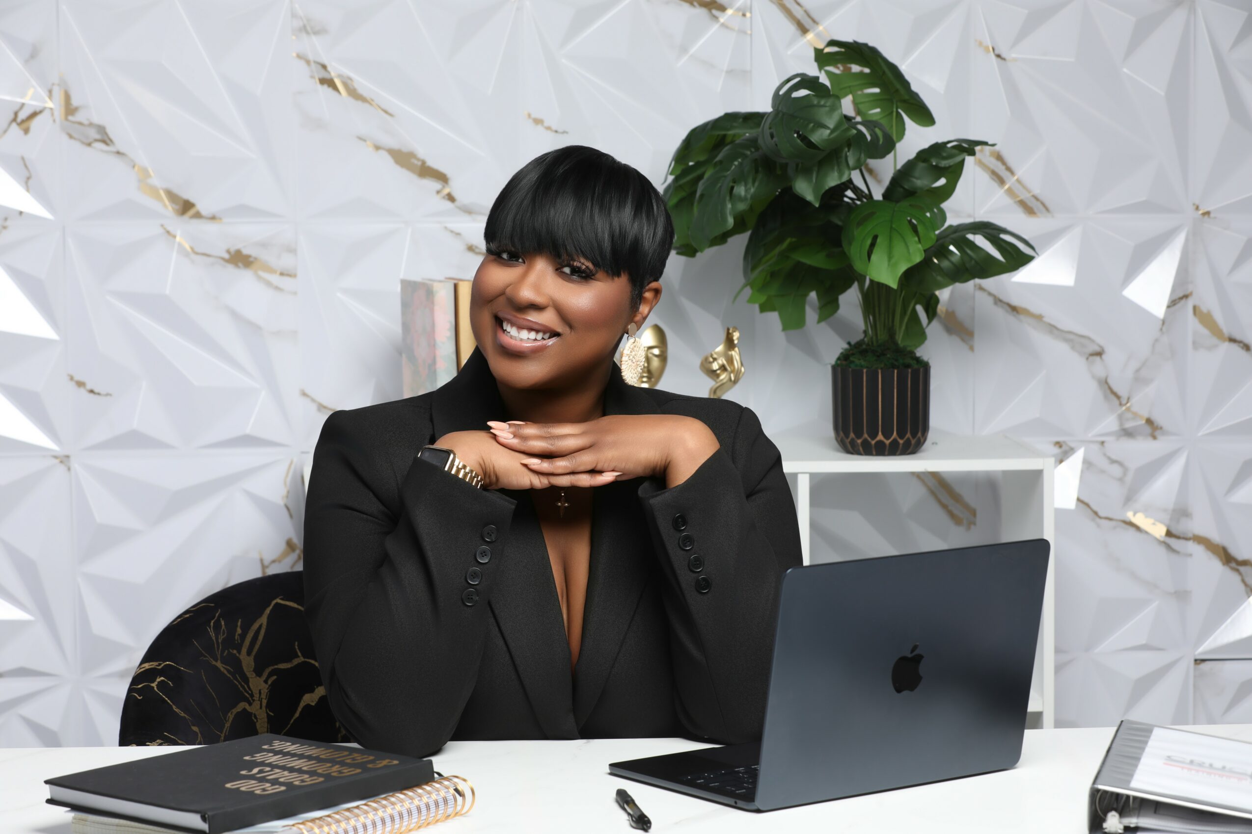 Woman with short black hair smiling, sitting at desk with laptop, in a modern room with white textured wall and green plant.