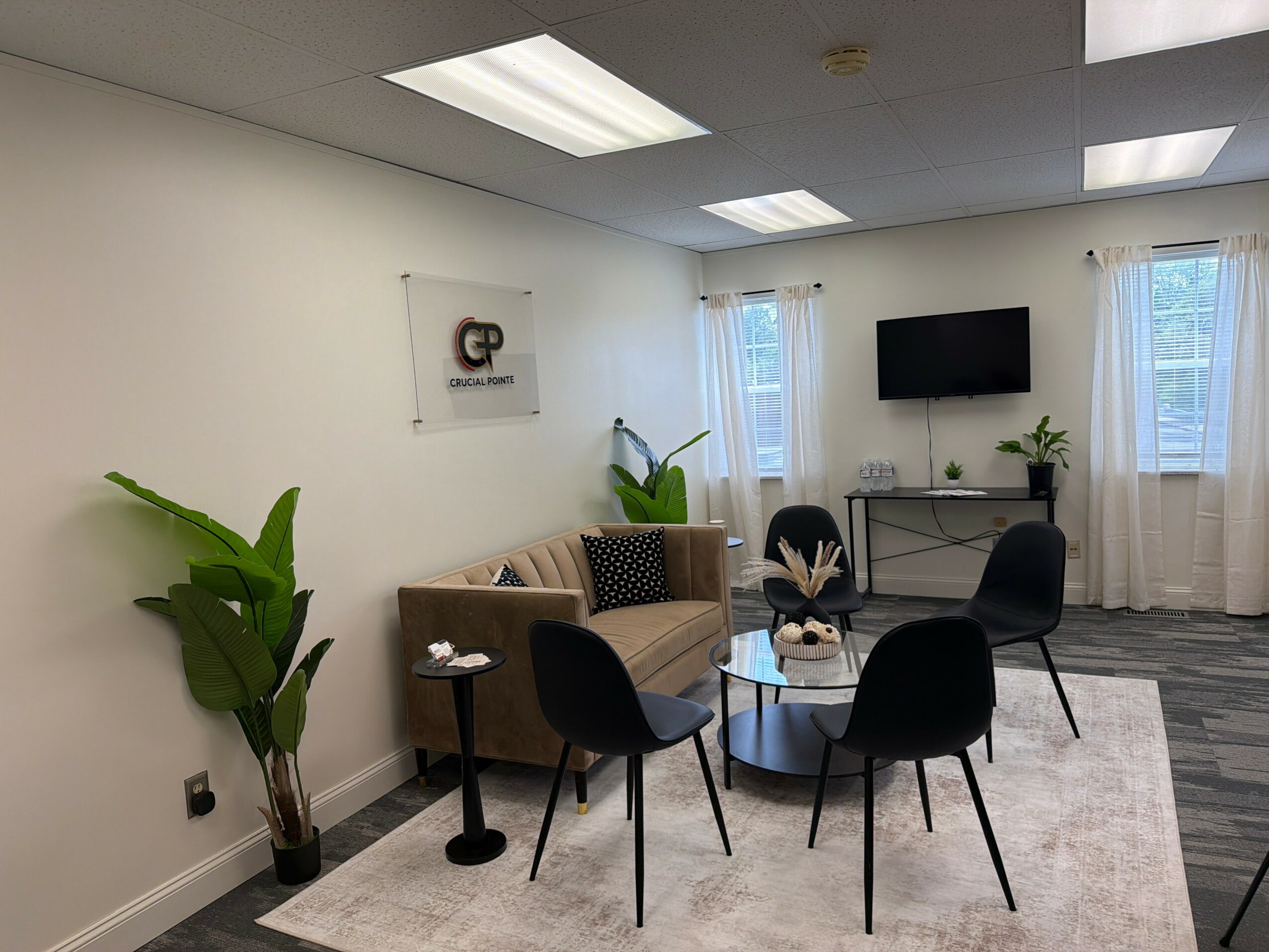 Office lounge with beige sofa, four black chairs around a glass table, potted plants, wall sign, and a wall-mounted TV.