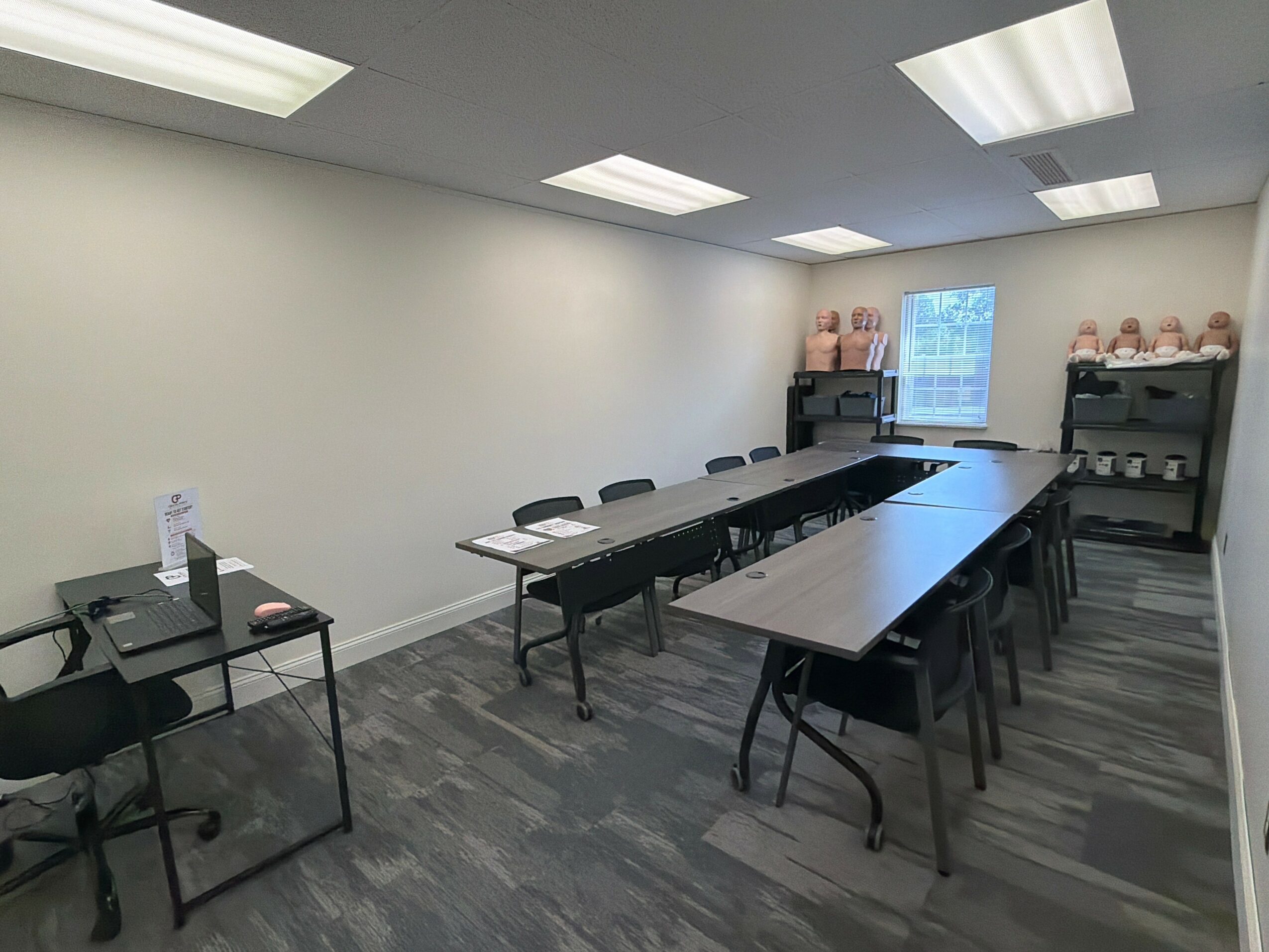 Empty conference room with long tables, chairs, shelves with pink objects, and a window, ceiling lights, and gray flooring.