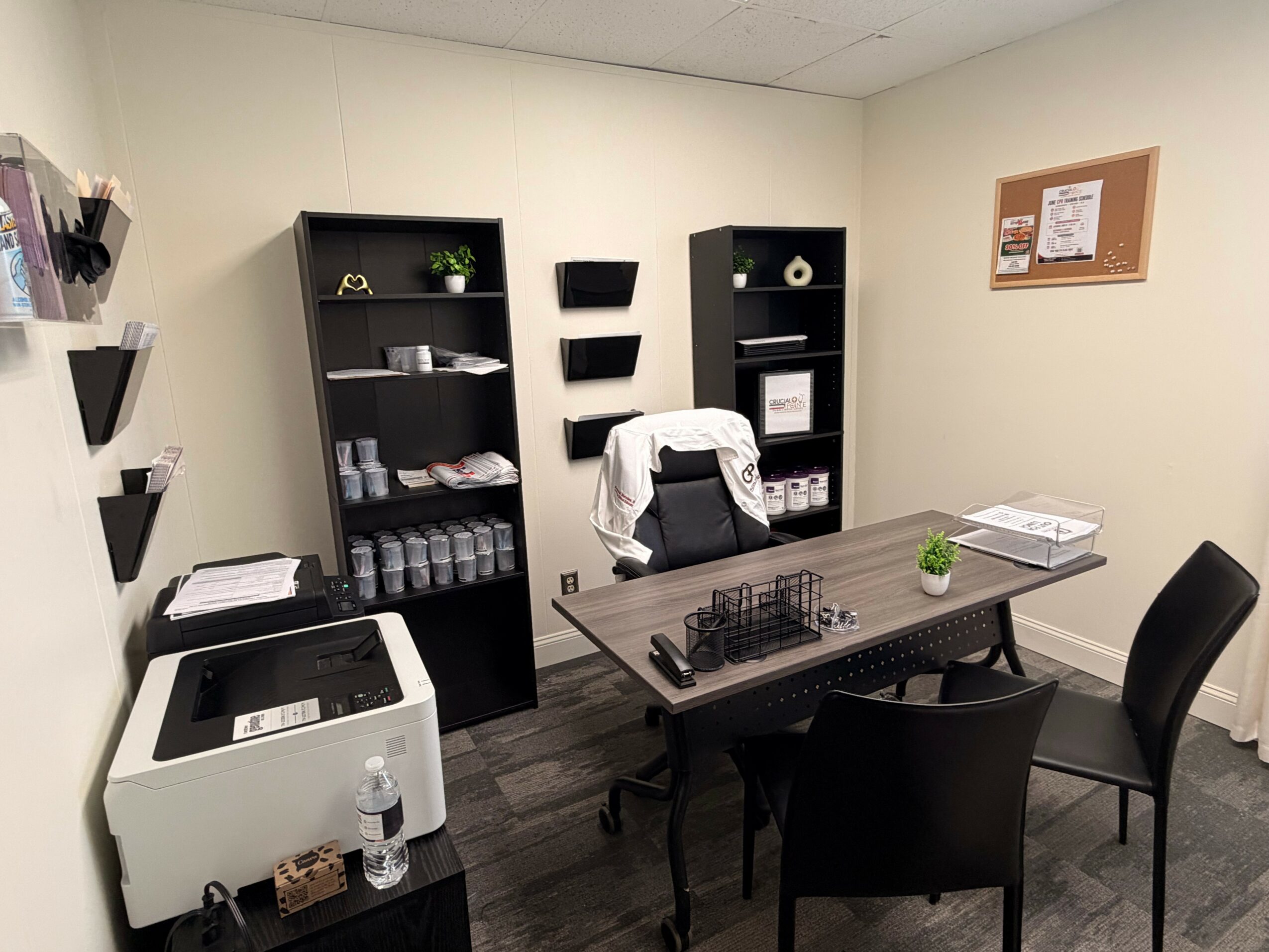 Office room with shelves, a desk, chairs, and a bulletin board on the wall.