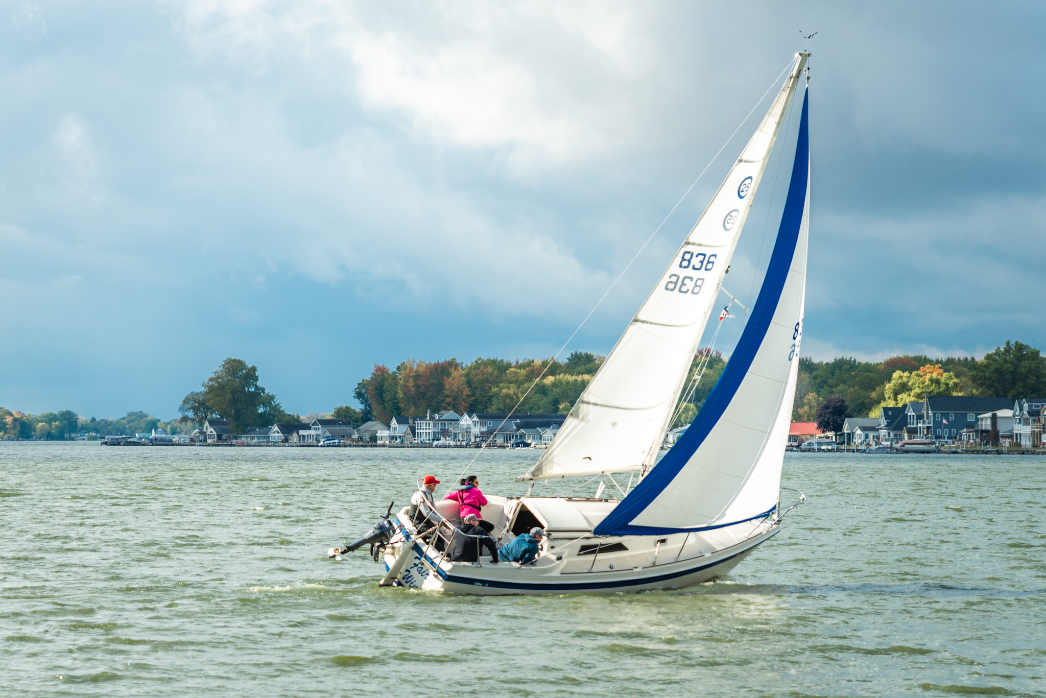 Sailboat with white and blue sails on water, with a shoreline and trees in the background.