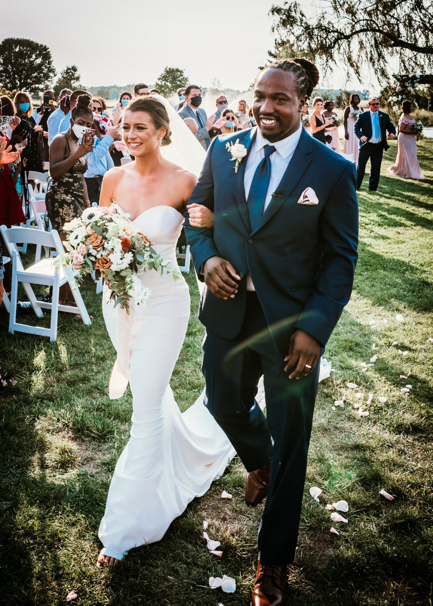 Bride and groom walking outdoors, smiling, surrounded by guests, trees, and chairs, during a wedding celebration.