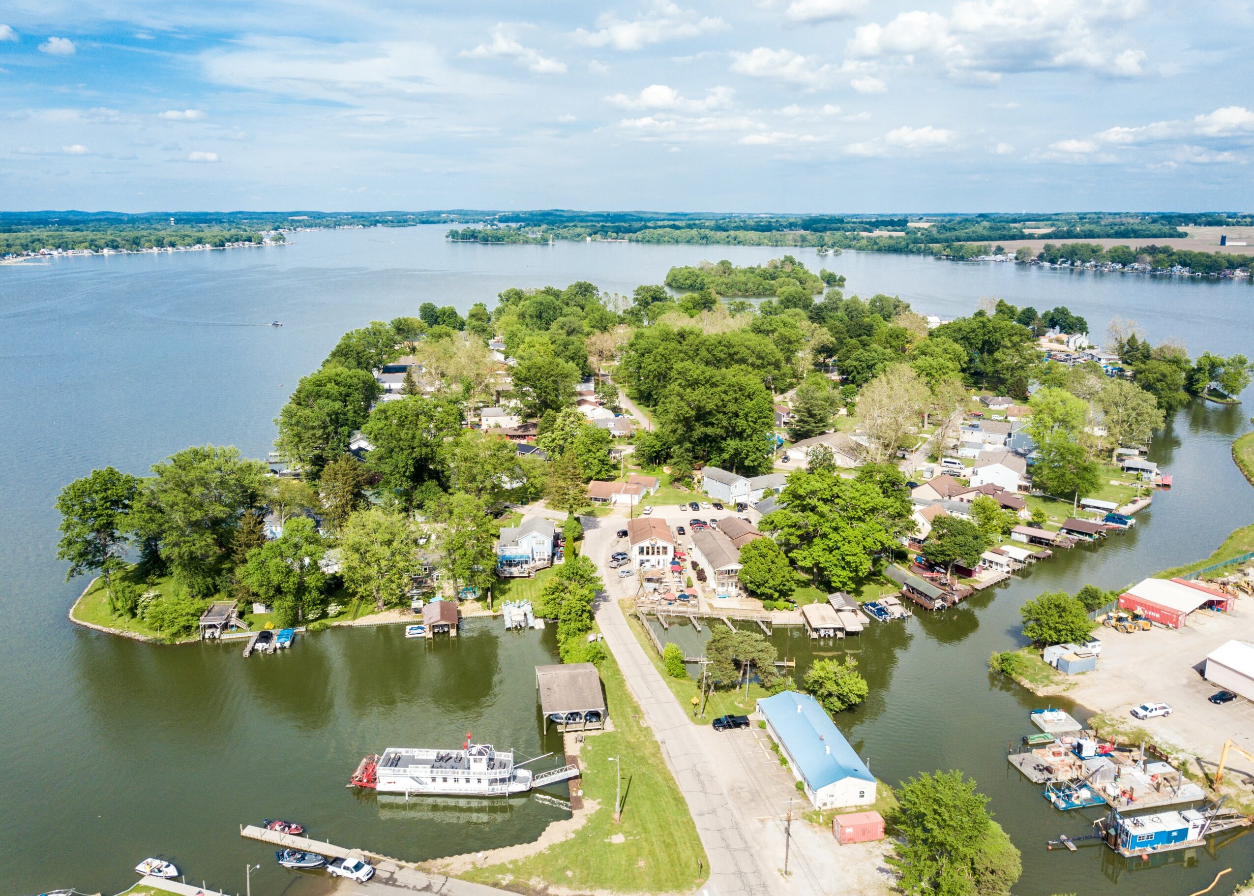Aerial view of a lakeside community with houses, trees, boats, and water, under a partly cloudy sky.