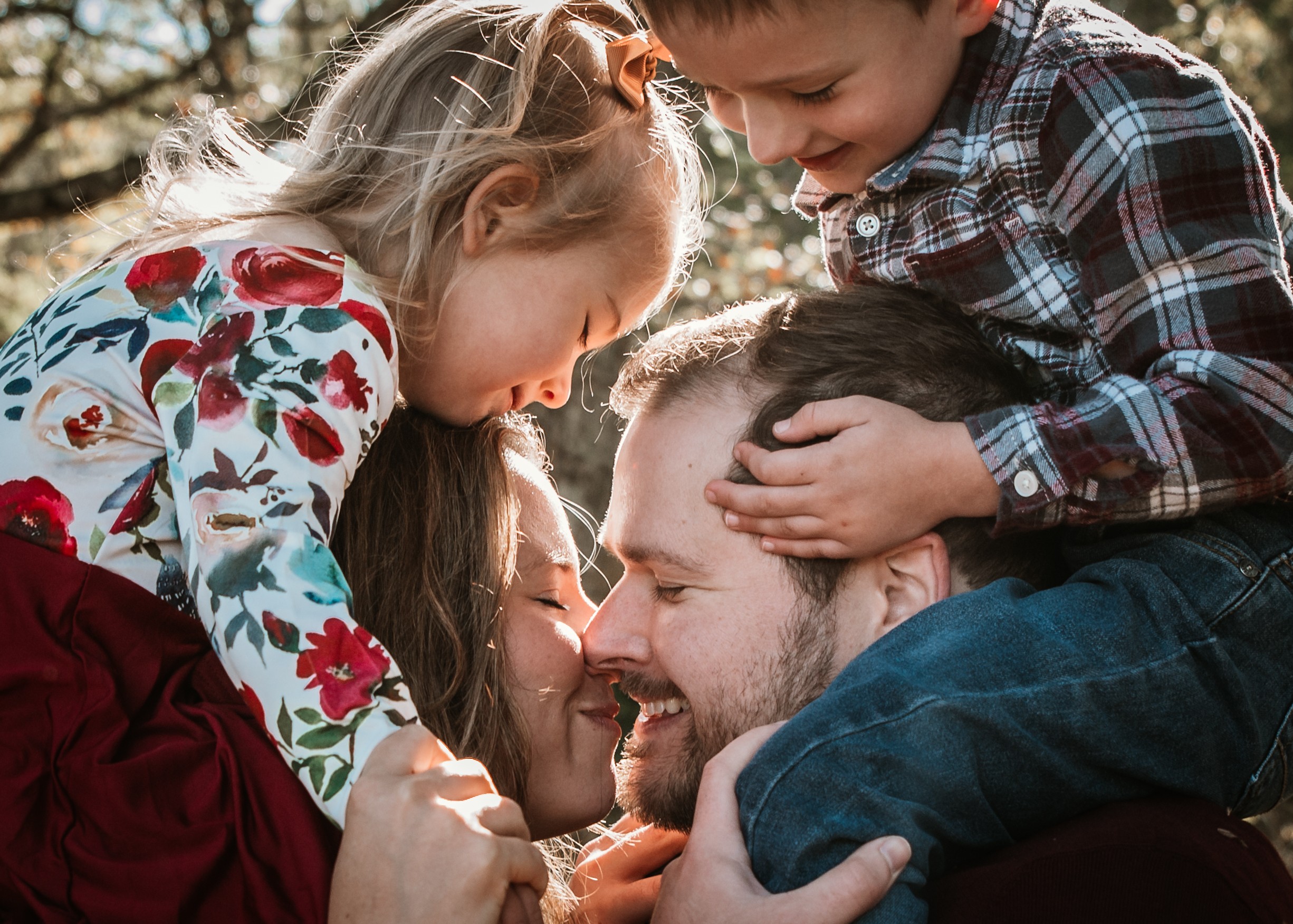 Family of four smiling and close together outdoors, children on parents' shoulders, sunlight filtering through trees.